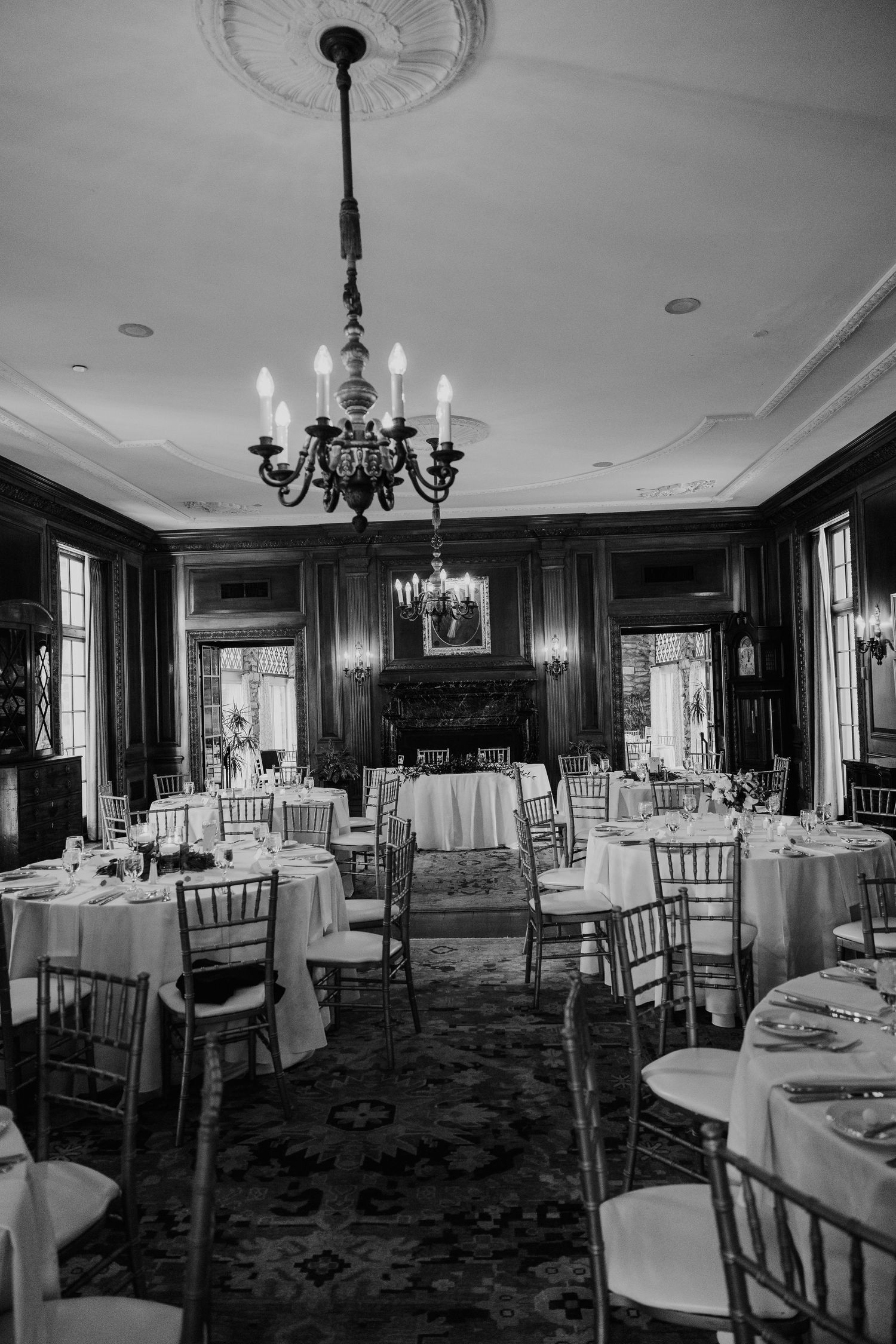 Elegant black and white photo of a wedding reception room with chandeliers, round tables, and chiavari chairs.