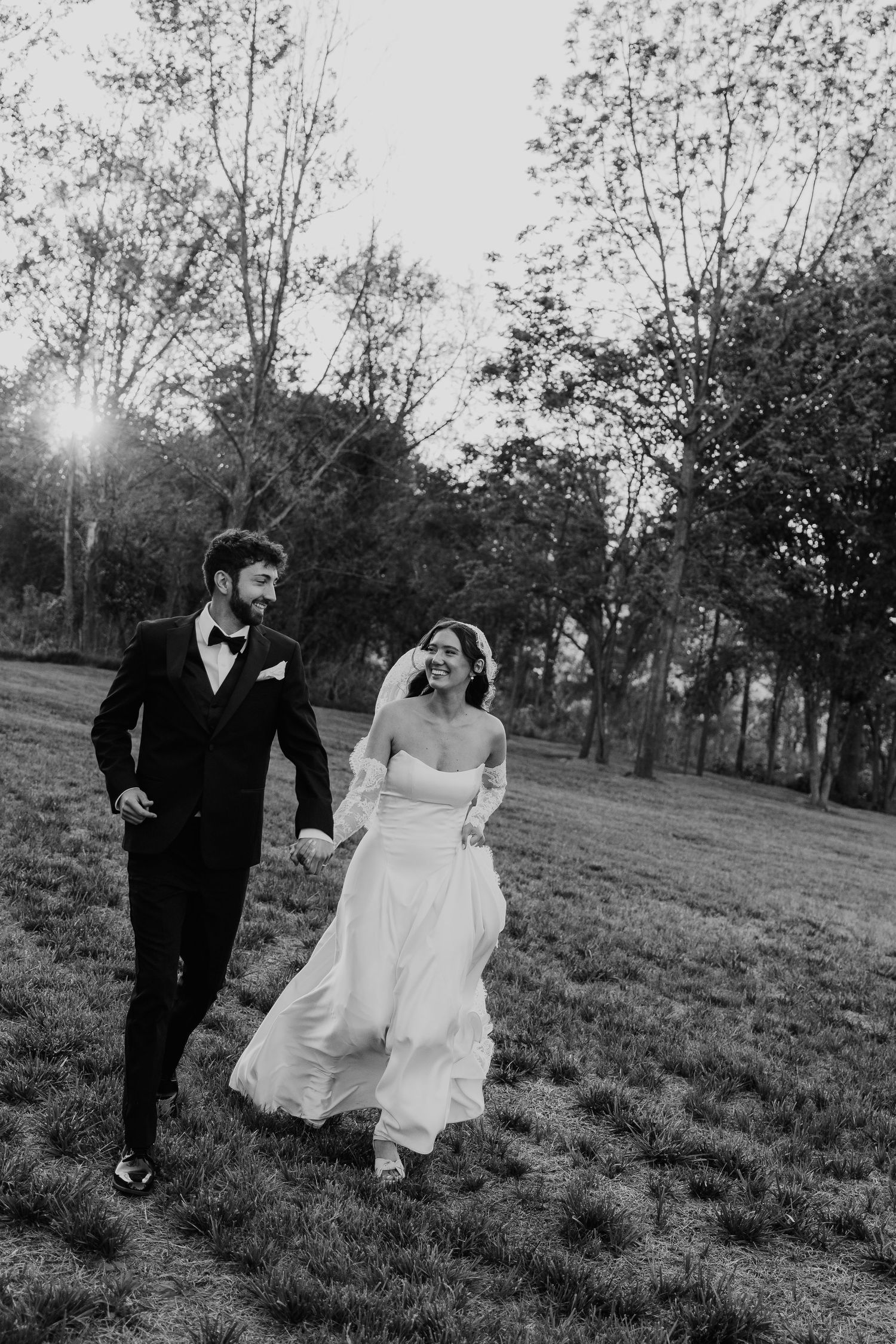 Black and white photo of smiling bride and groom holding hands walking through a sunlit garden on their wedding day.