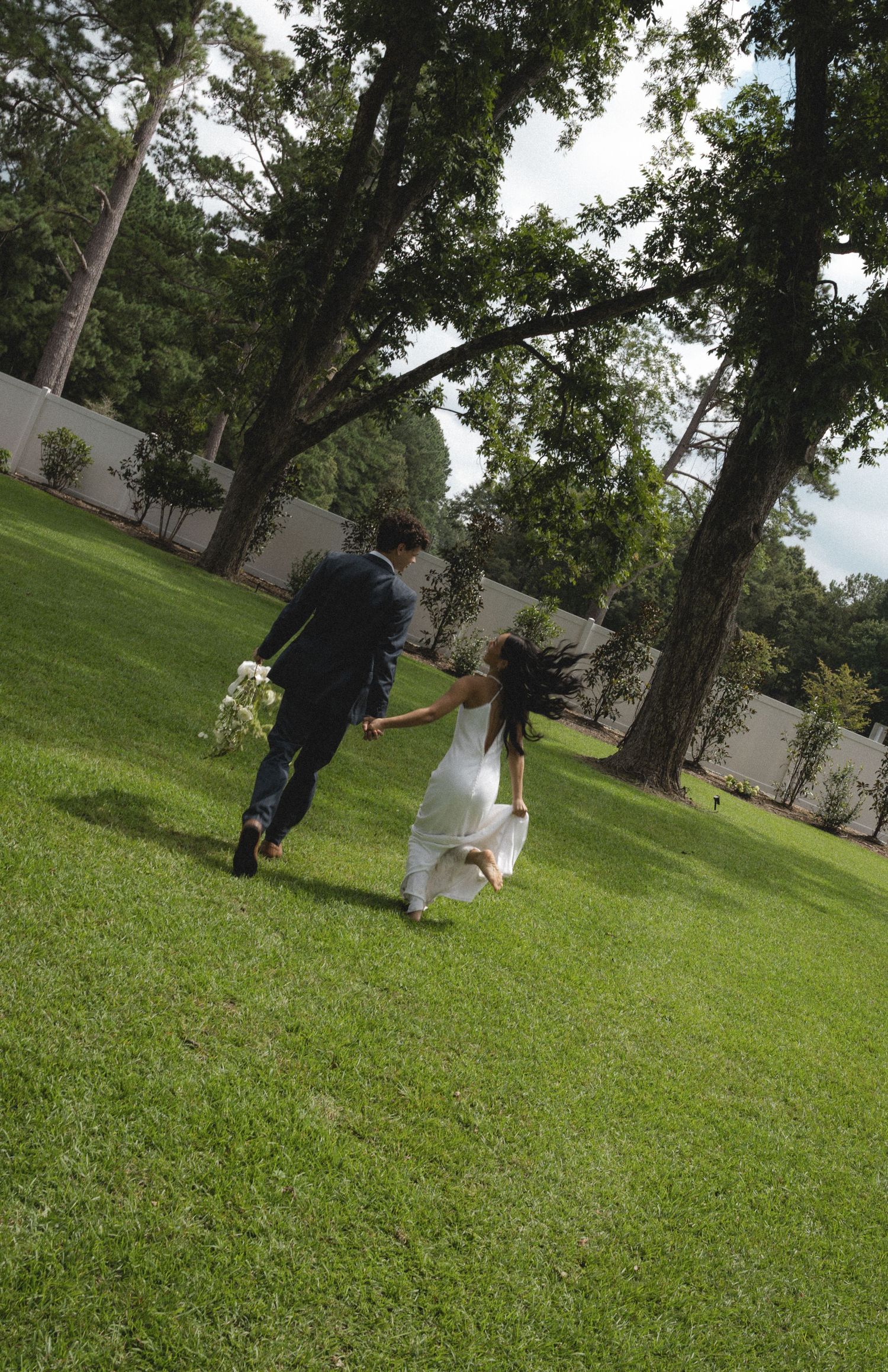 Couple runs playfully across green lawn surrounded by tall trees.