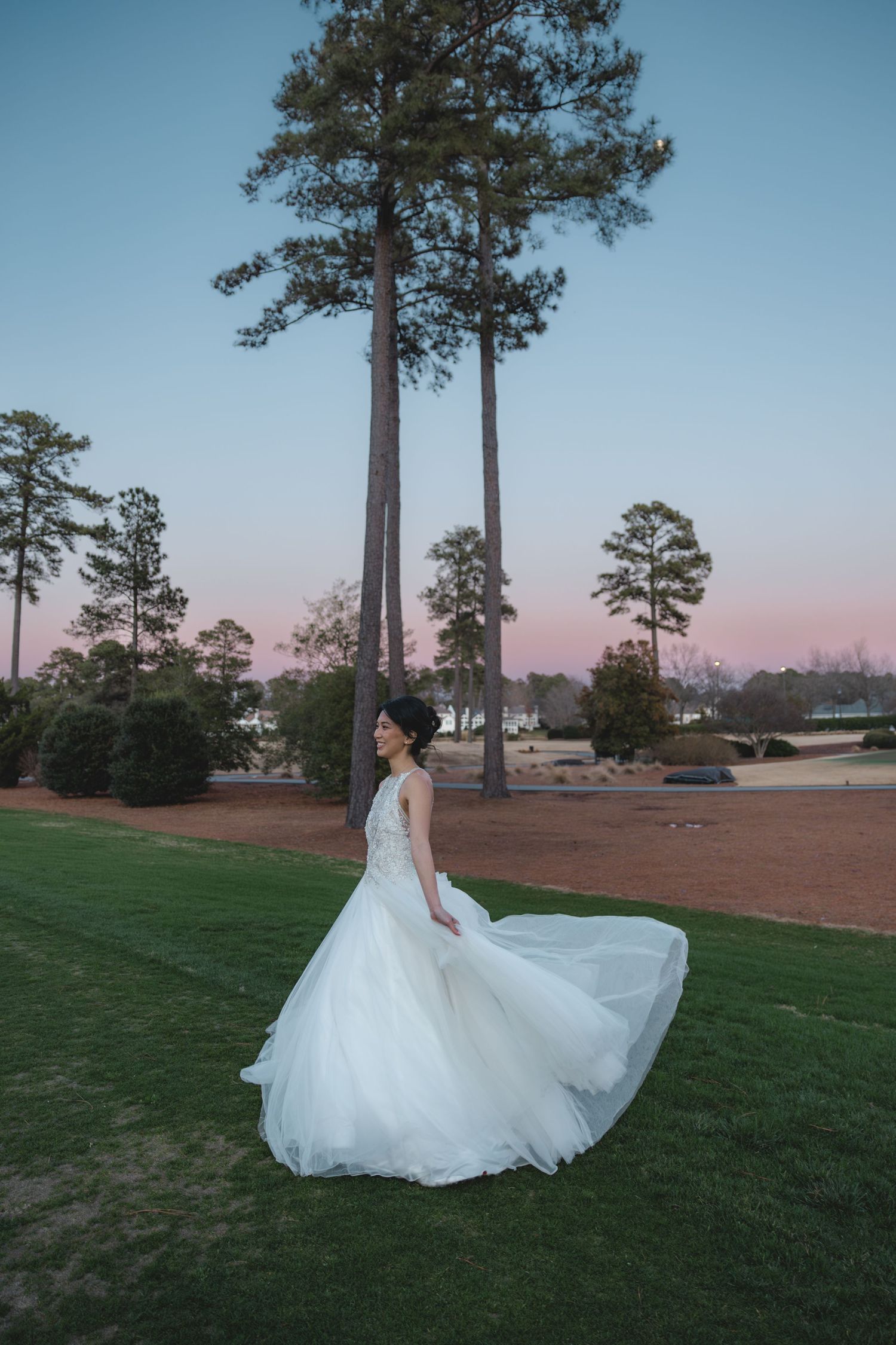 Bride in flowing white ball gown twirling at sunset beside tall pine trees.