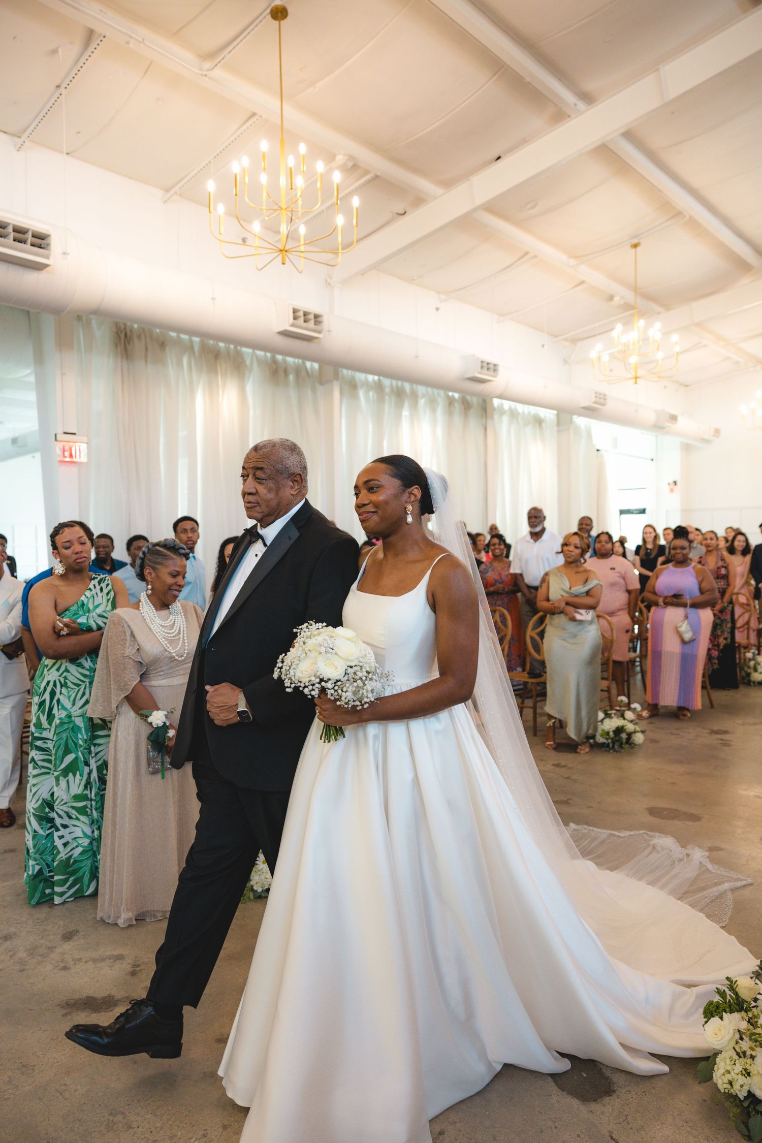 Bride in white ball gown walks down aisle with escort as wedding guests look on in bright venue.