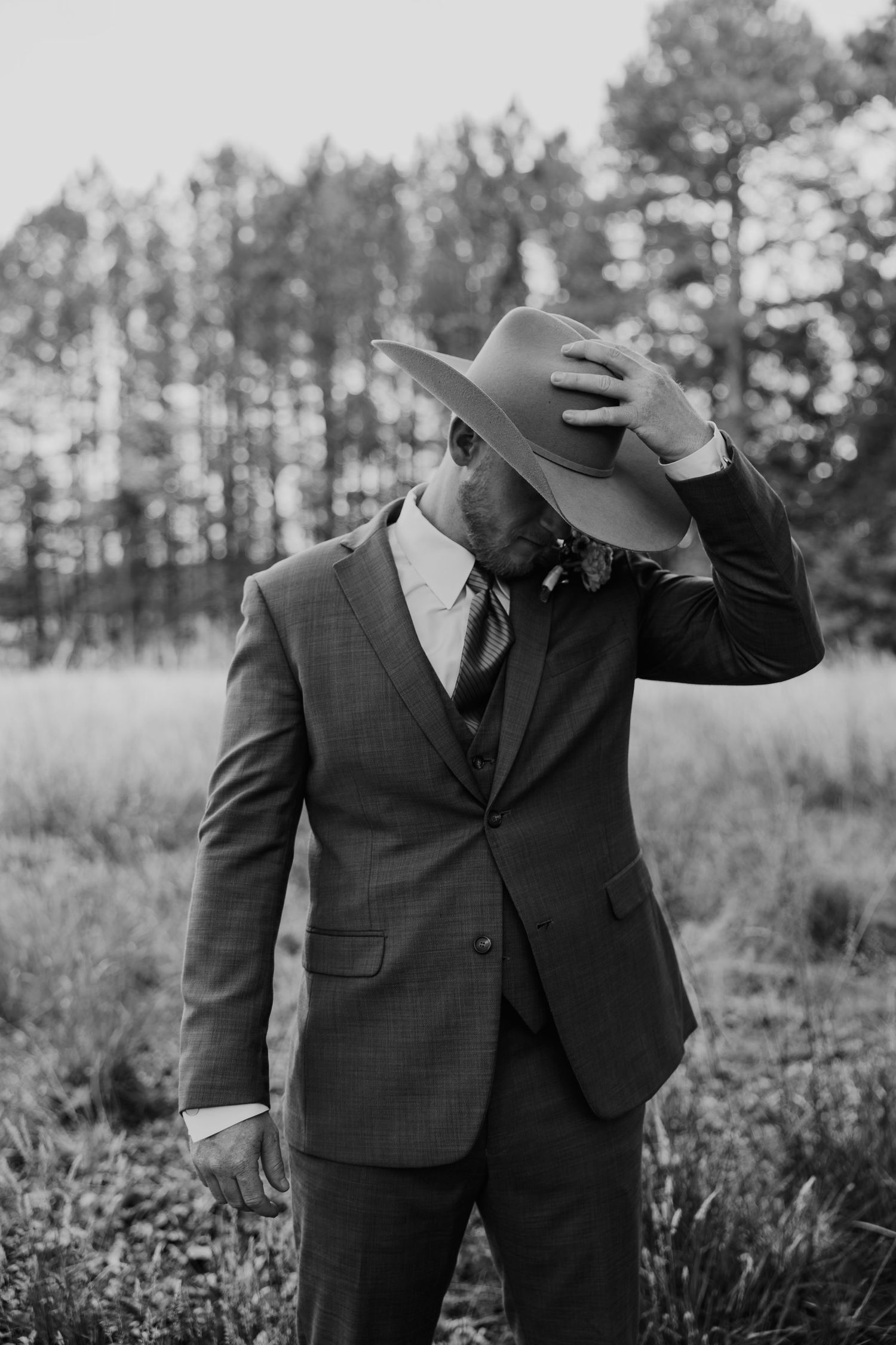 Black and white portrait of groom in suit tipping cowboy hat in pine forest.