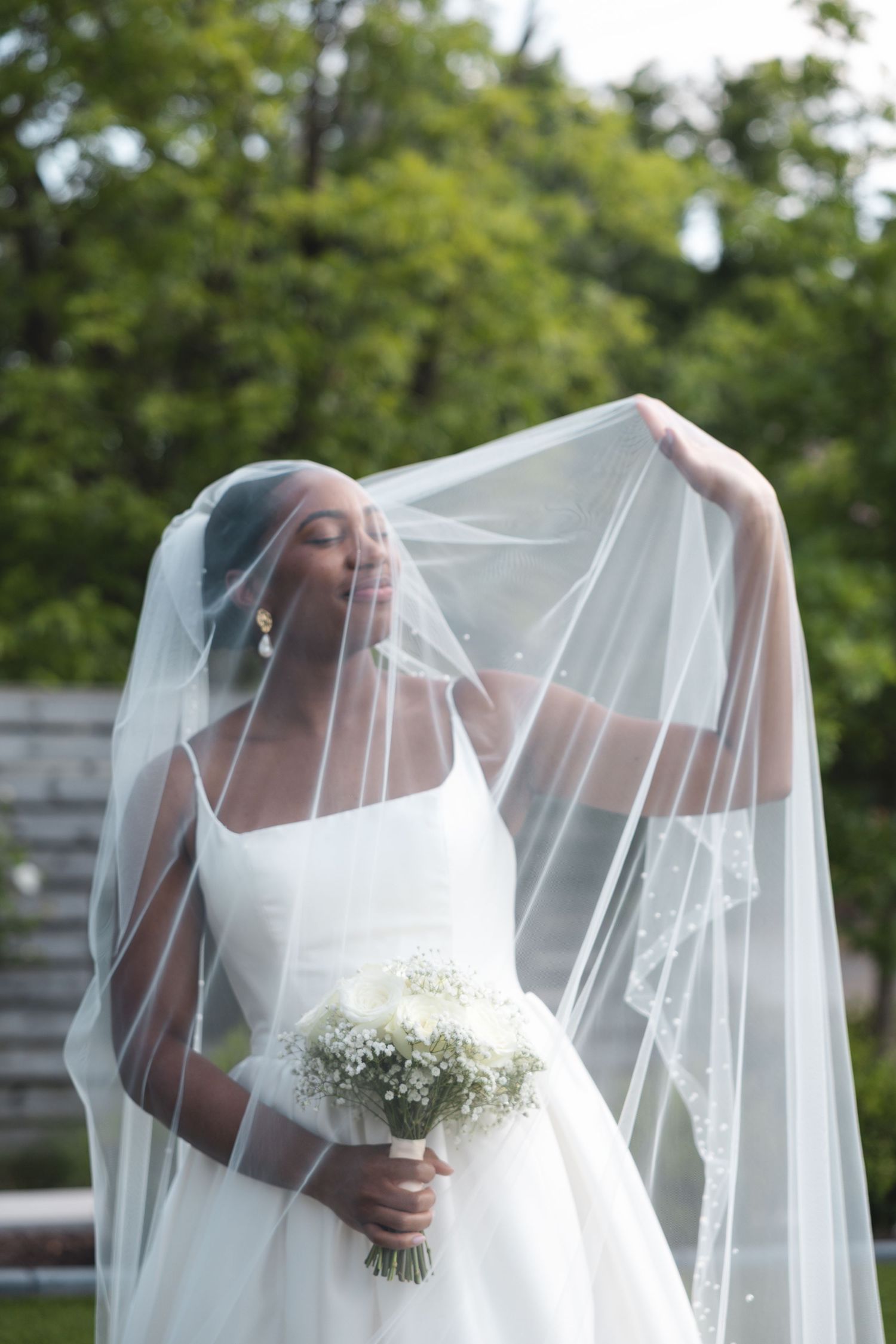 Elegant bride in white gown lifts her lace veil while holding a white bouquet outdoors.