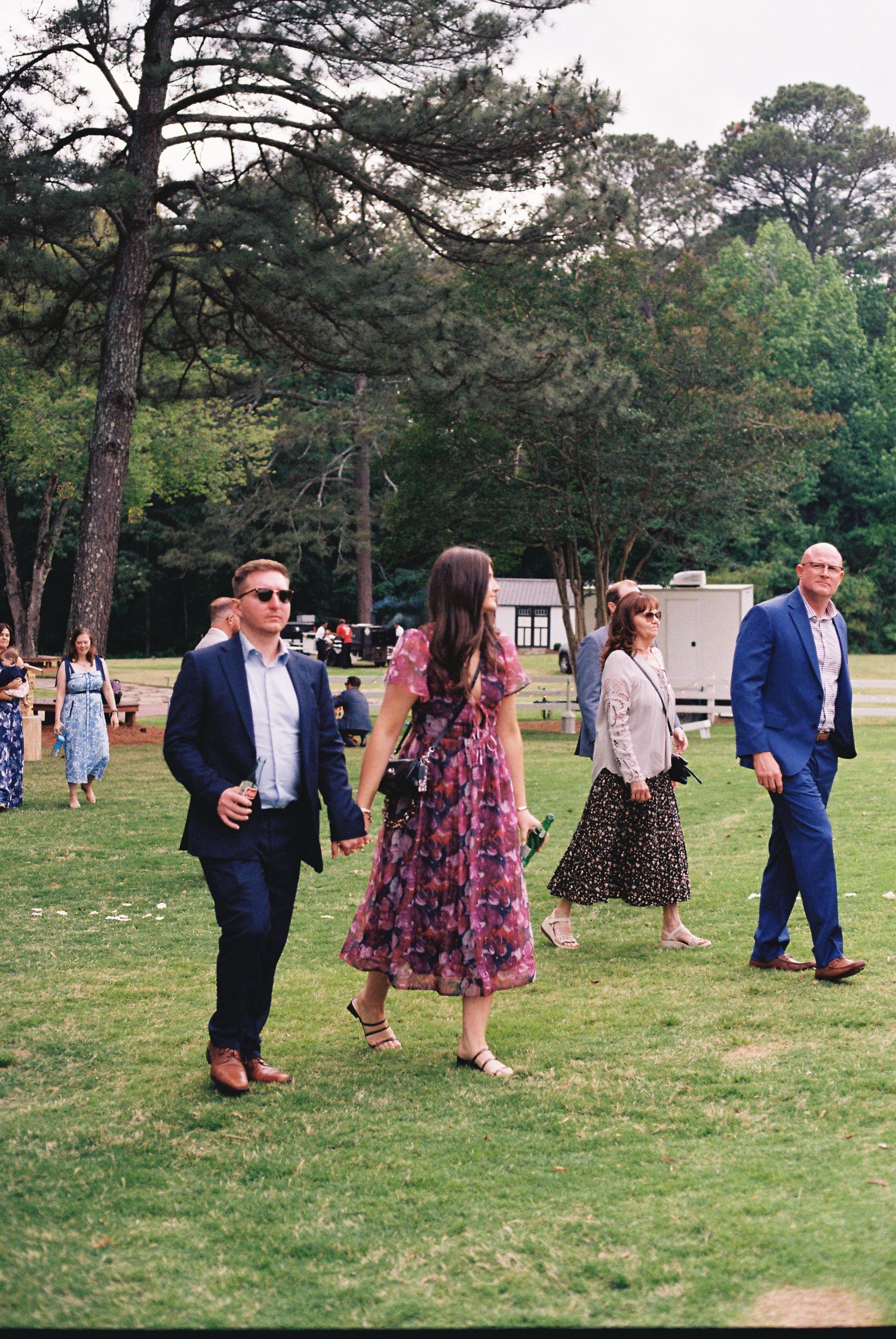 Elegantly dressed wedding guests walking on green lawn surrounded by tall pine trees at an outdoor garden event.