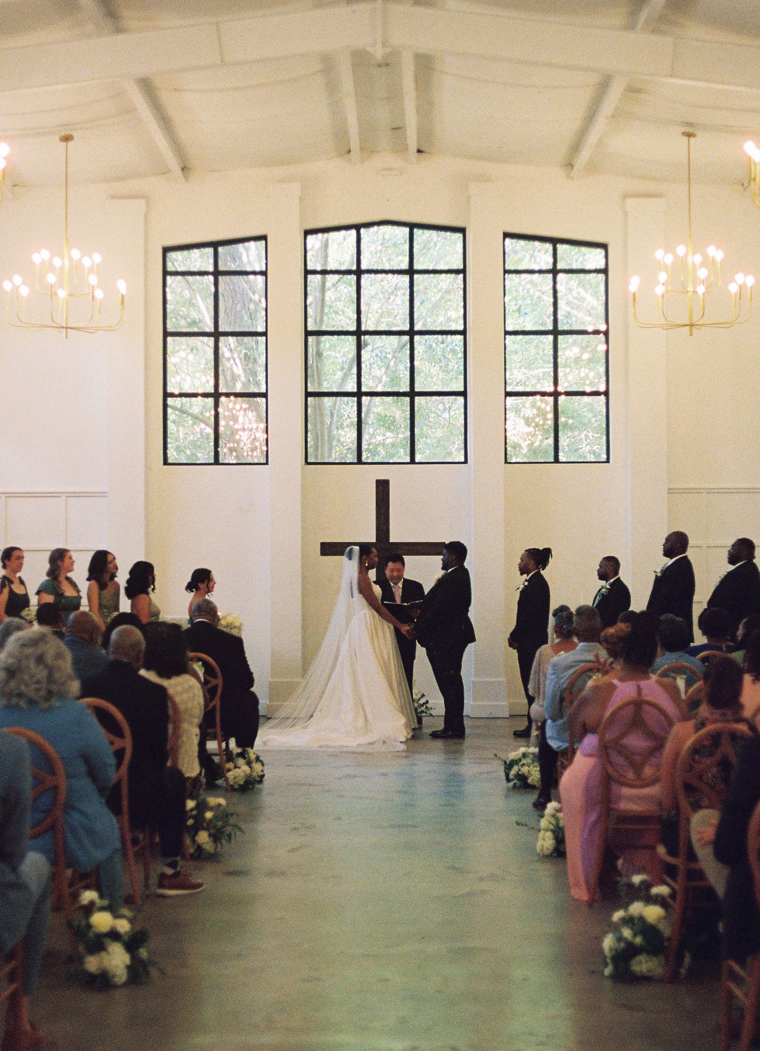 Elegant wedding ceremony in a white chapel with large windows, bride and groom at altar with wooden cross, guests seated in wooden pews.