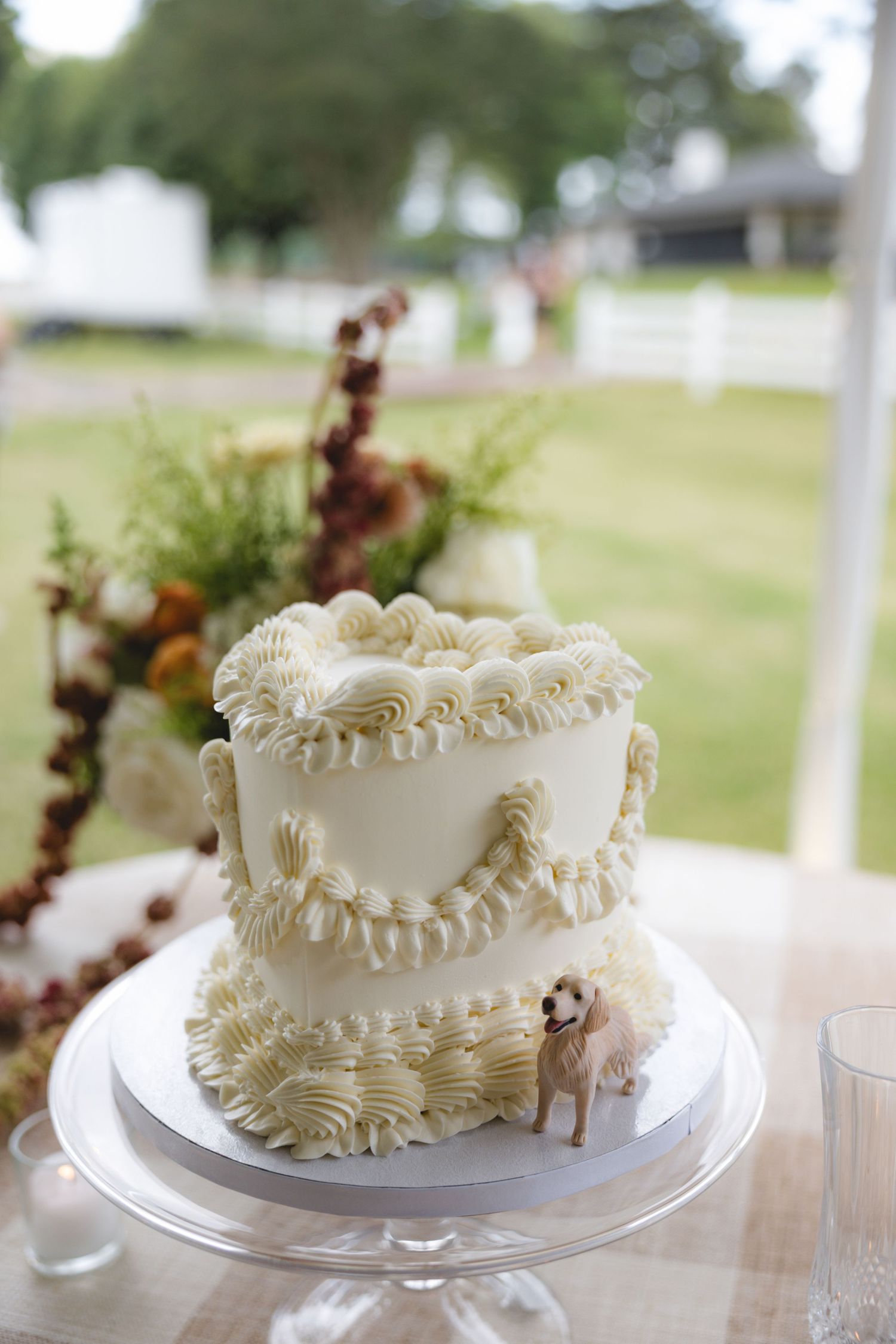 Two-tier white wedding cake with intricate piped frosting details and a small dog figurine topper on a glass cake stand outdoors.