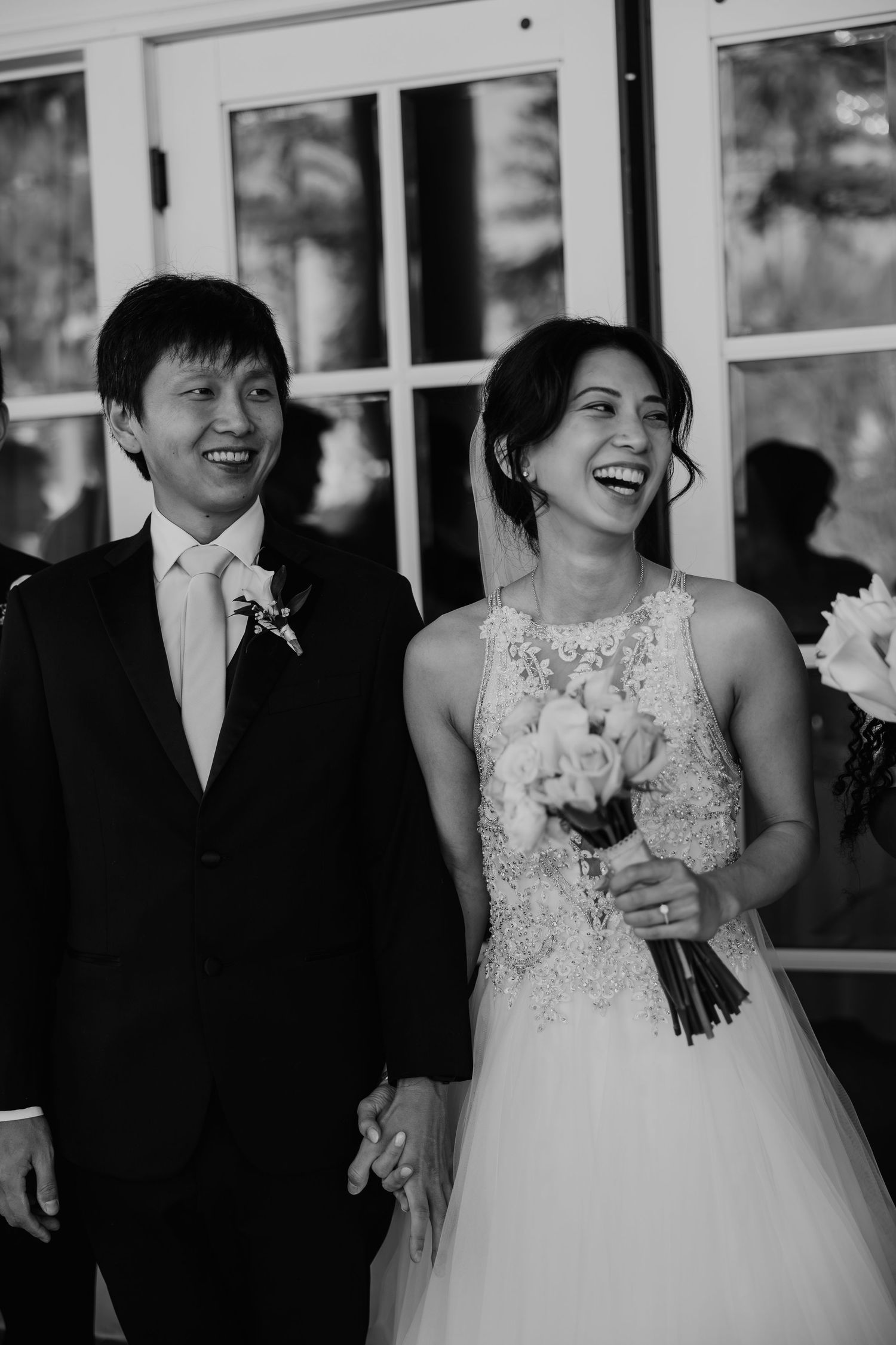 Black and white photo of a smiling bride holding flowers and groom in a tuxedo at their wedding ceremony.