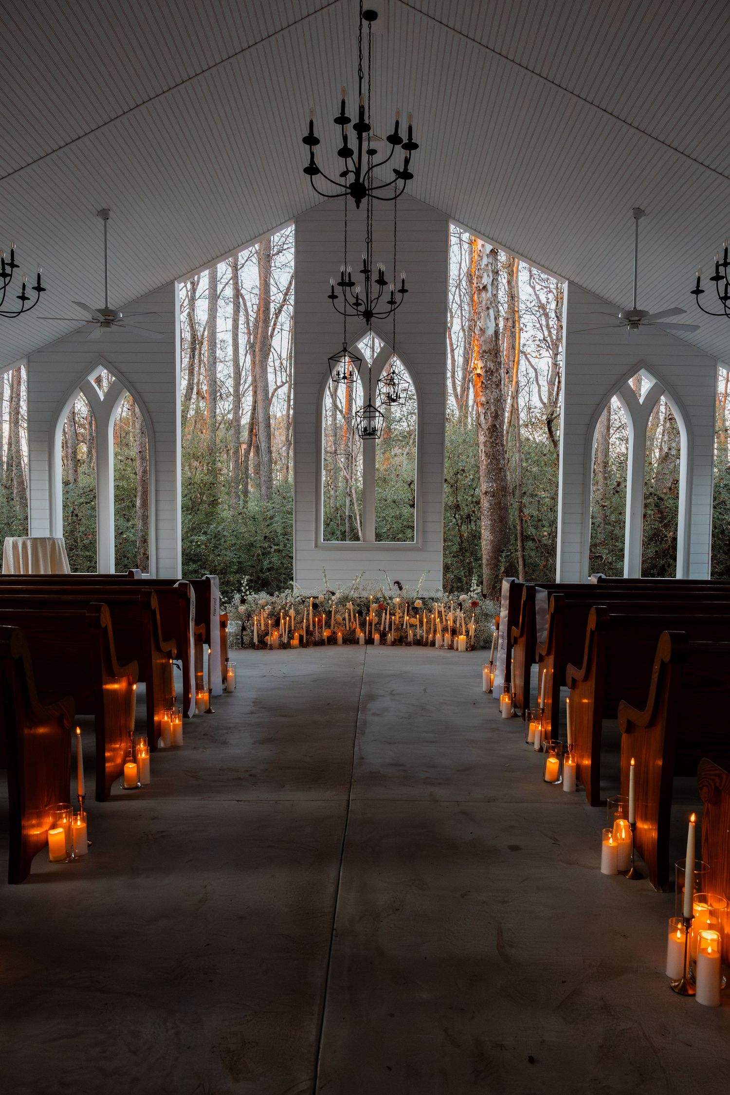 Candlelit chapel interior with wooden pews, gothic arched windows, and chandeliers creating a romantic wedding ceremony ambiance.