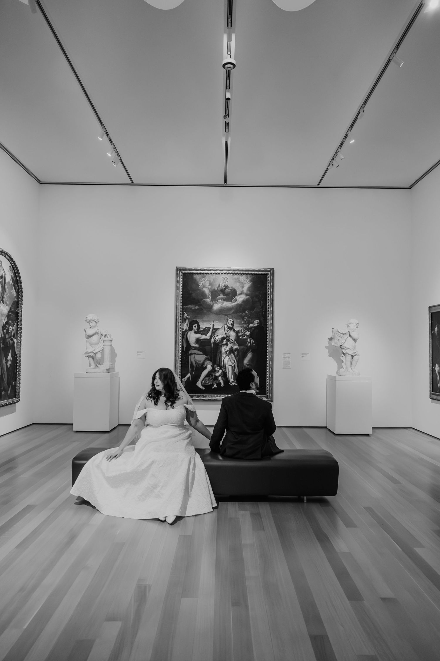 Black and white photo of a bride and groom sitting on a bench in an art museum gallery with classical paintings and sculptures.
