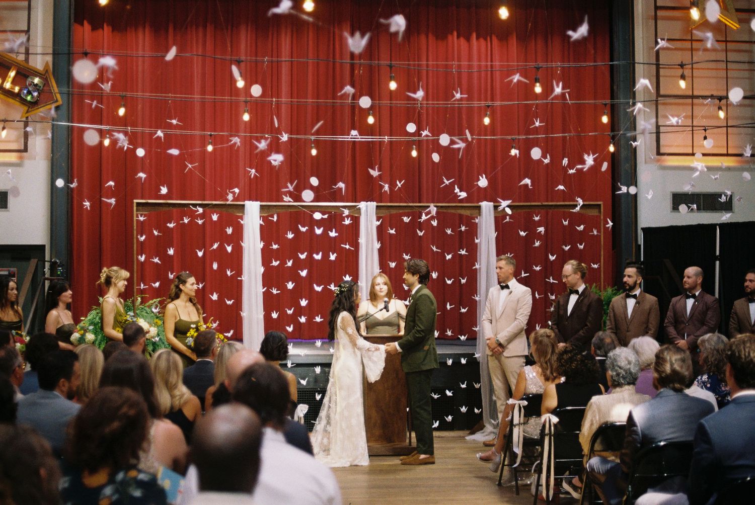Wedding ceremony with paper cranes falling from above, couple at altar before red curtain backdrop.