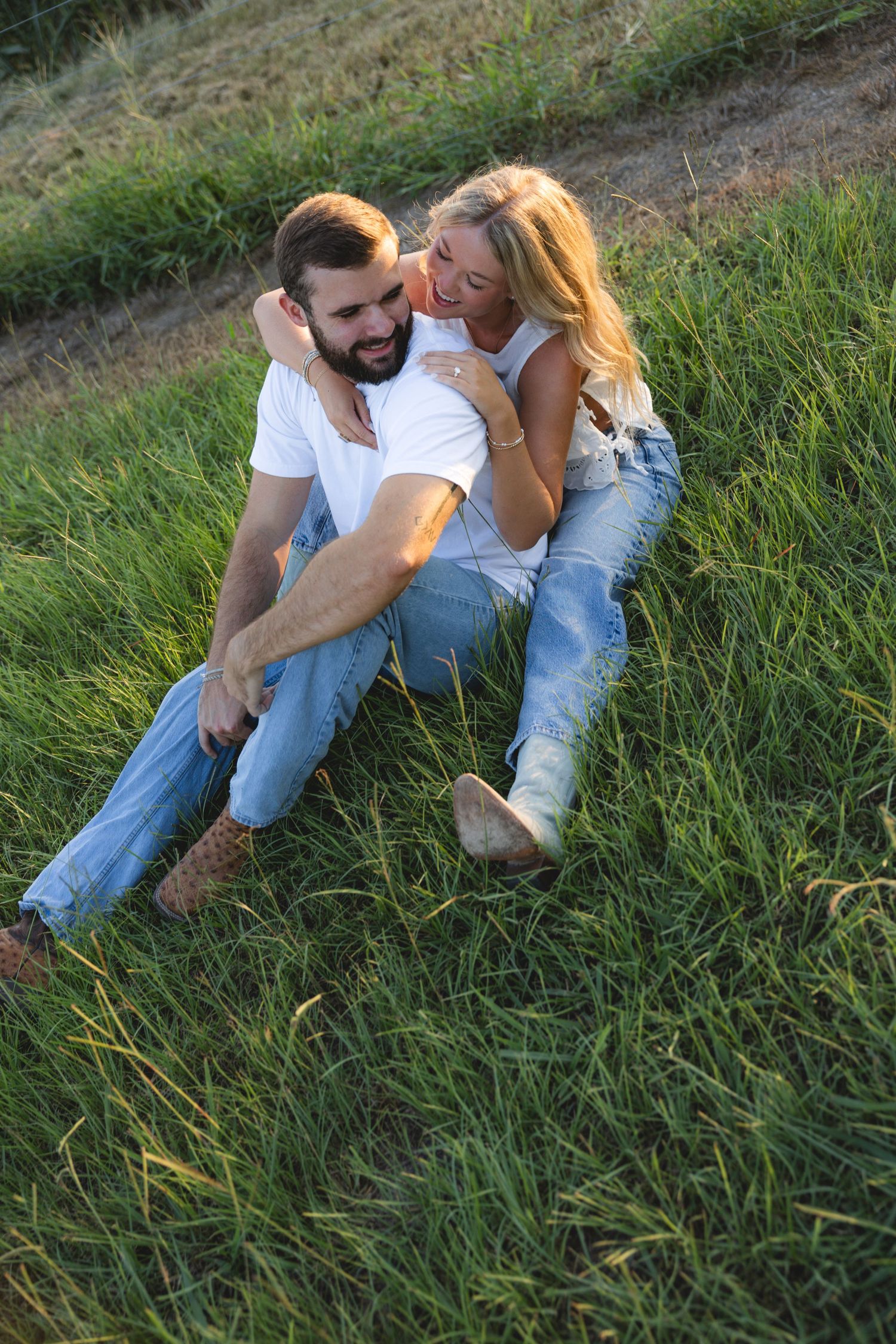 Happy couple sitting together in green grass field, woman embracing man from behind, both wearing jeans and white tops.