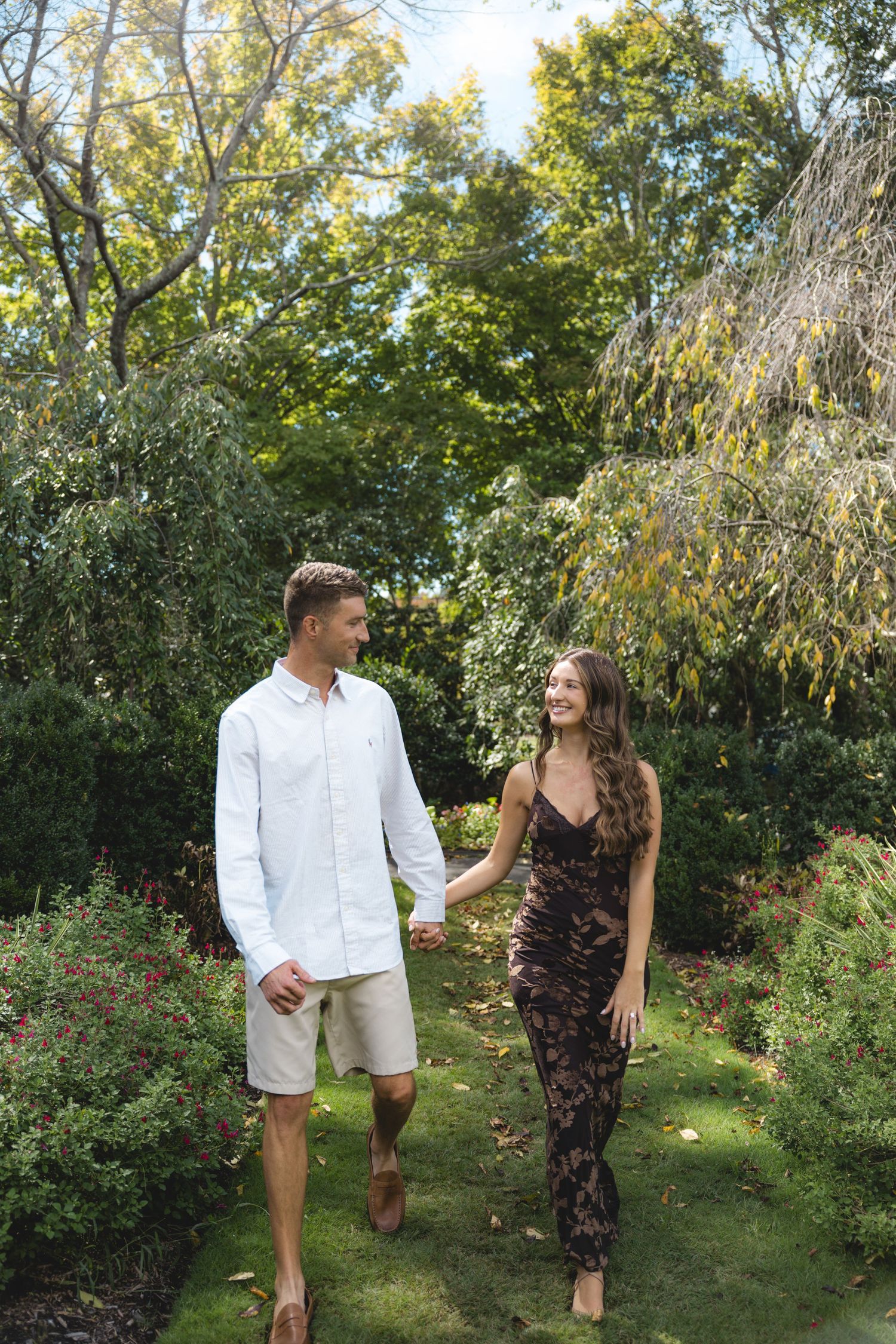 A couple holds hands and smiles while walking through a lush garden, man in white shirt and woman in floral dress.