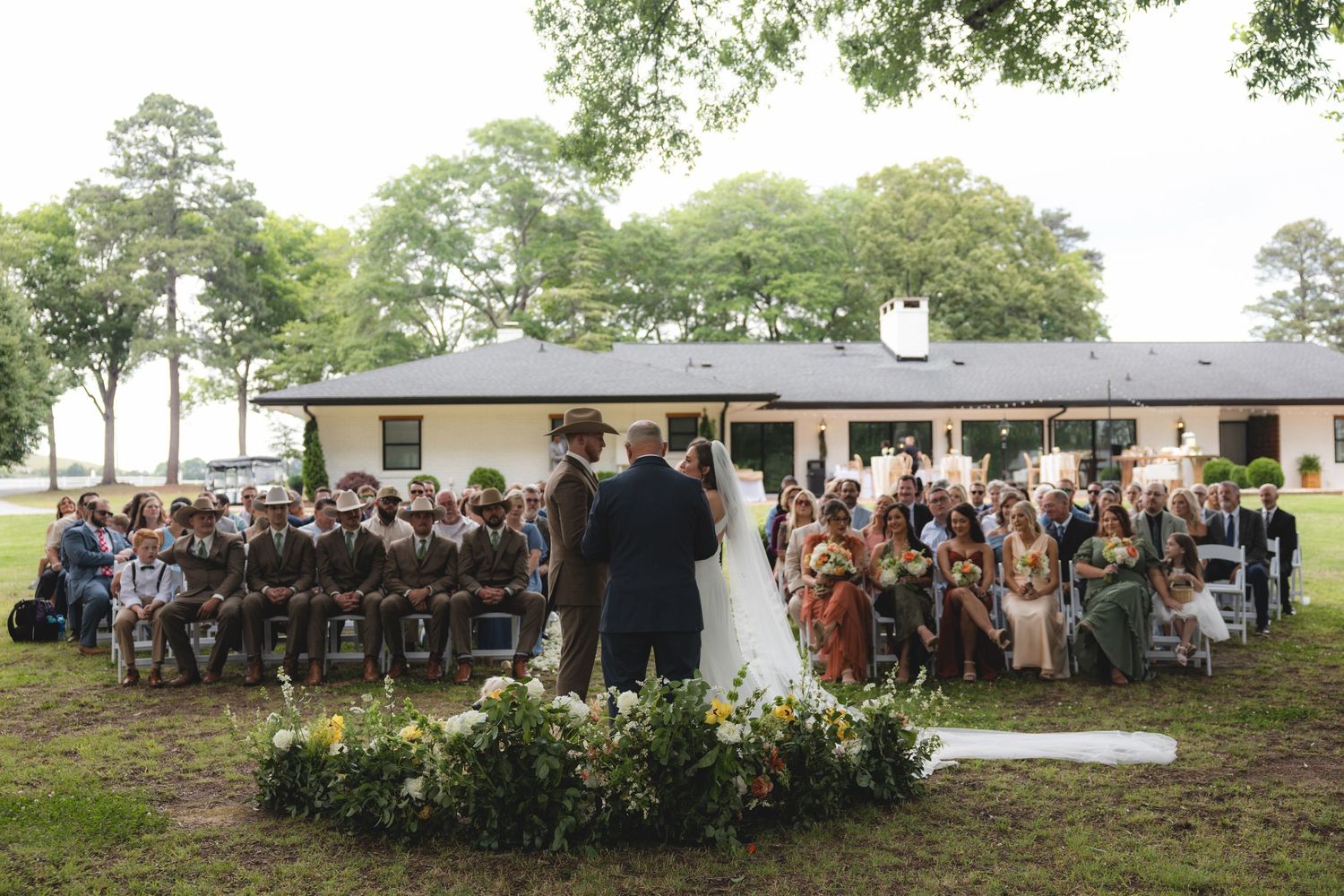 Aerial view of outdoor wedding ceremony with guests seated in rows facing bride and groom at altar.