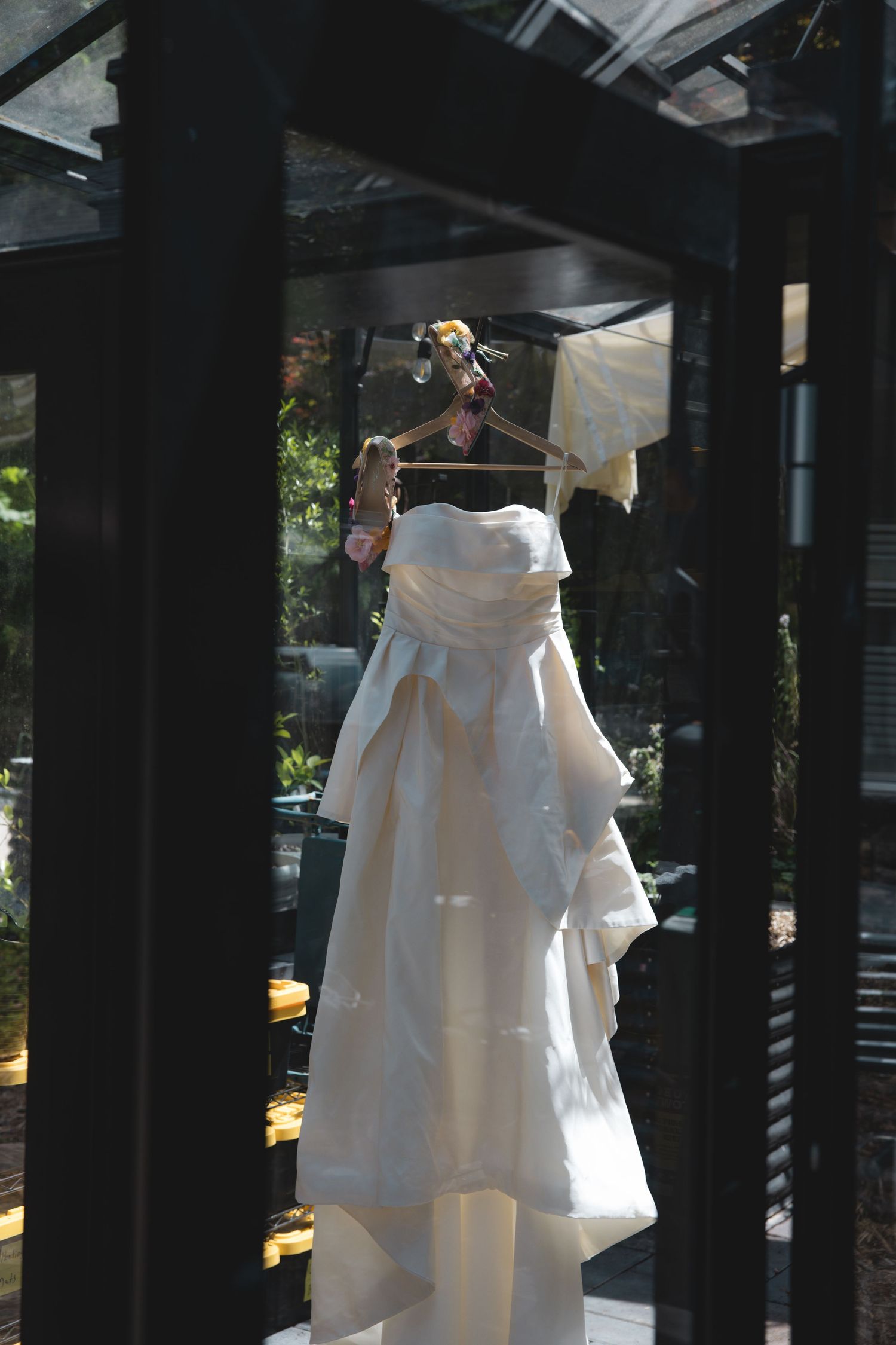 Elegant strapless white wedding dress hanging on decorative hanger in sunlit greenhouse setting.