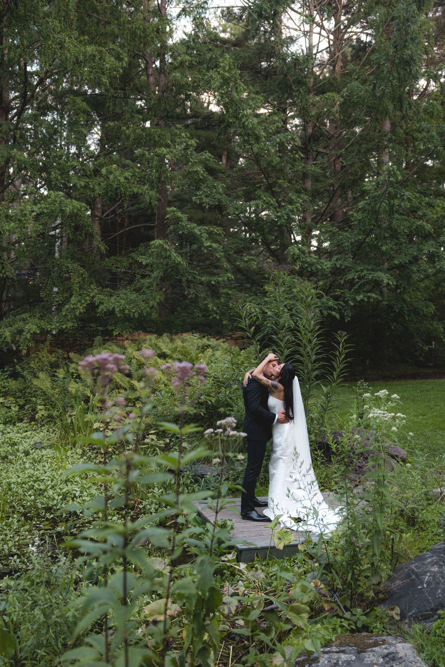 Bride and groom embracing on a wooden bridge surrounded by lush green forest and wildflowers during outdoor wedding.