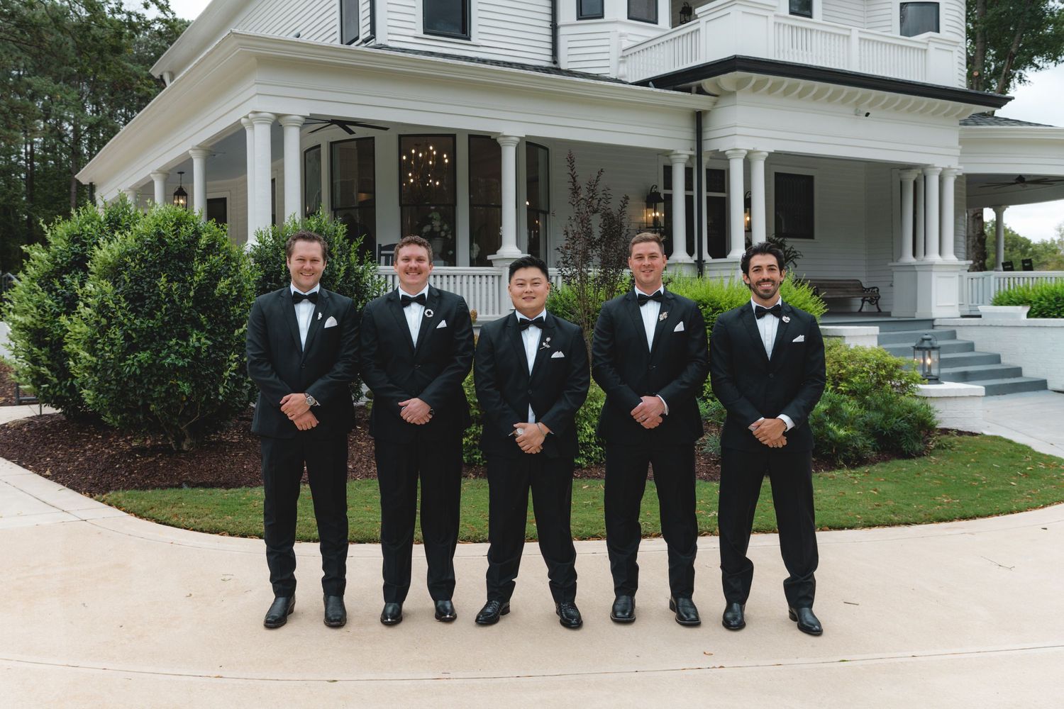 Five groomsmen in black tuxedos and bow ties stand in a row on a stone path in front of a white colonial-style venue with manicured shrubs.