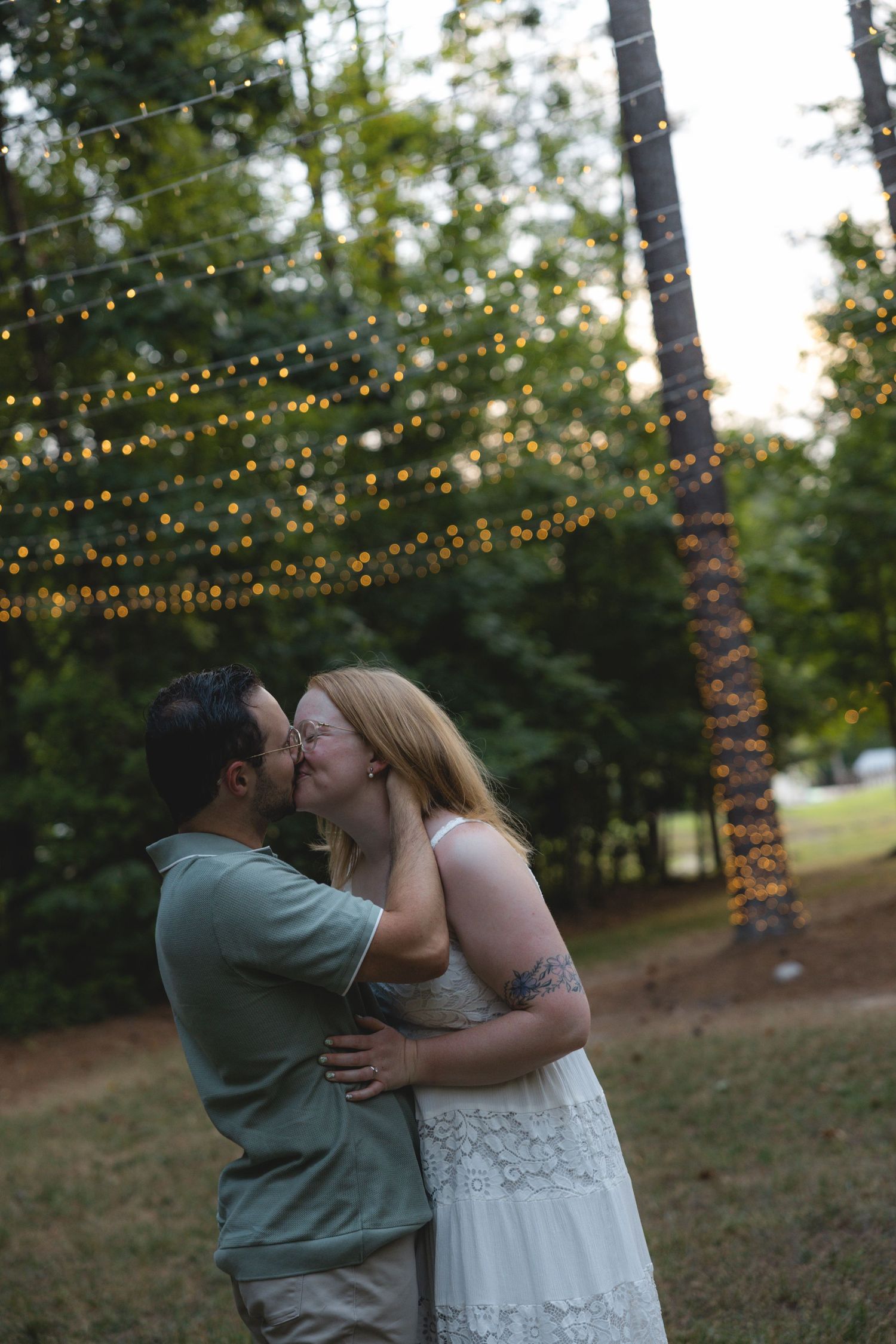 Couple sharing a romantic kiss outdoors under glowing string lights with lush green trees in the background at dusk.