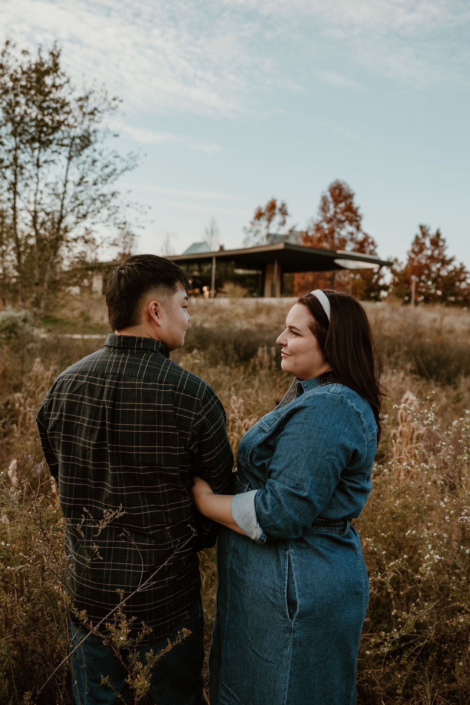 Couple sharing a romantic moment in an autumn field, woman in denim dress smiling at man in plaid shirt.