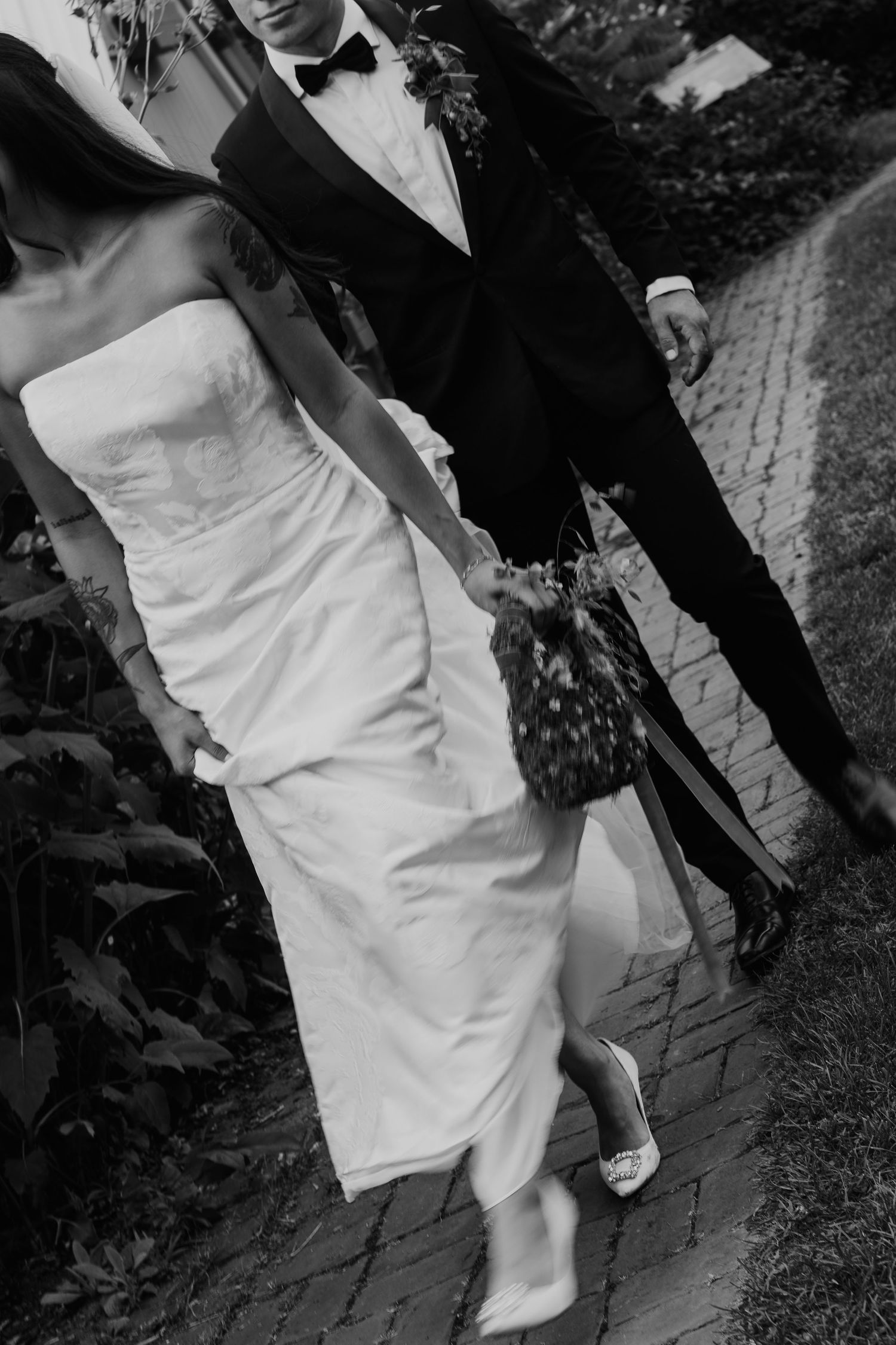 Black and white photo of a bride in a white gown and groom in a tuxedo walking hand in hand on a brick path.