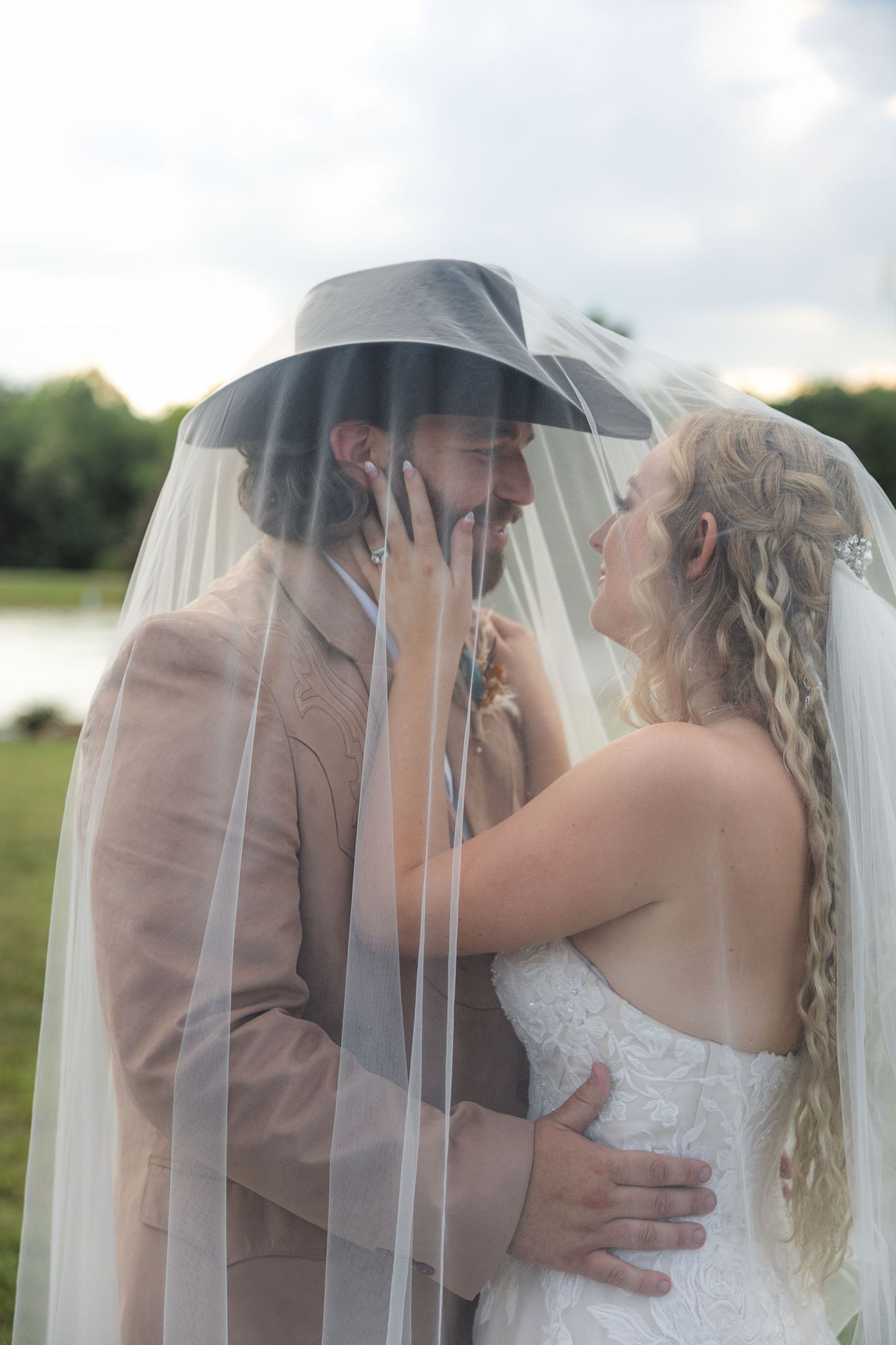 Bride and groom share a romantic moment under a wedding veil outdoors, groom wearing a cowboy hat and tan suit.