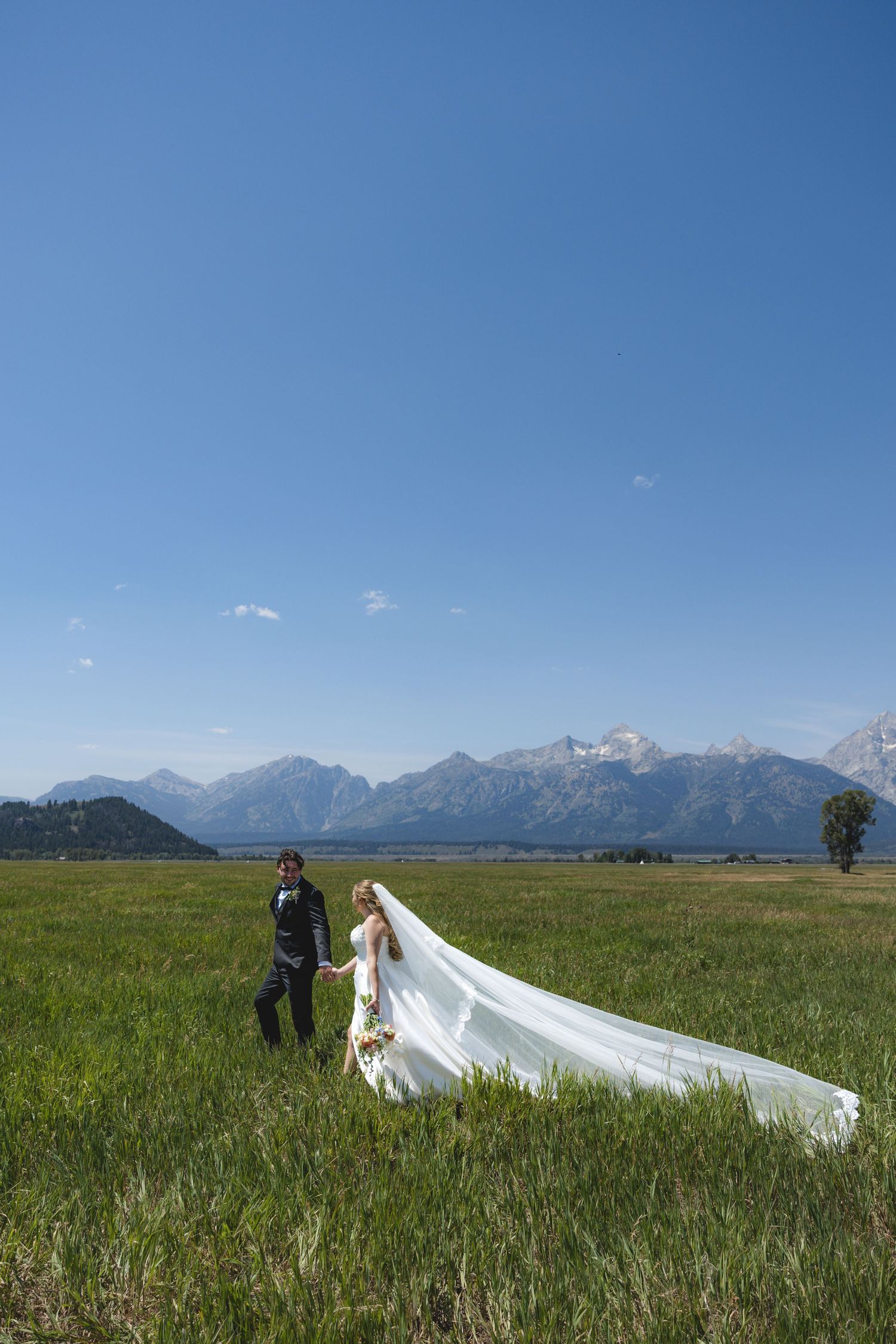 Bride and groom holding hands in green meadow with Grand Teton mountains backdrop, bride's veil flowing in wind.