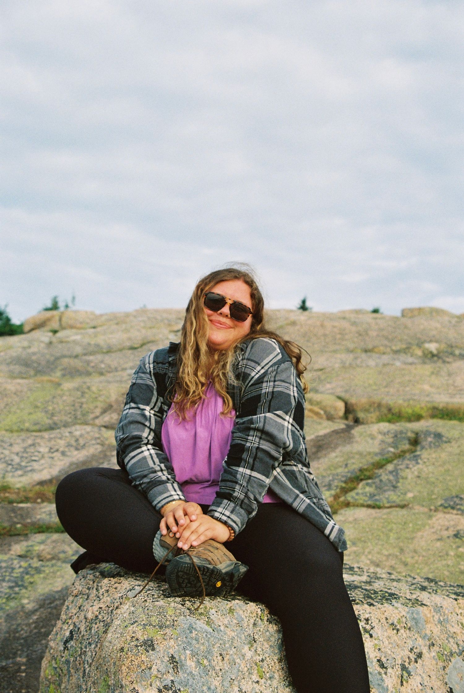 Woman in plaid jacket and sunglasses sitting on rocky terrain outdoors, smiling at camera on cloudy day.