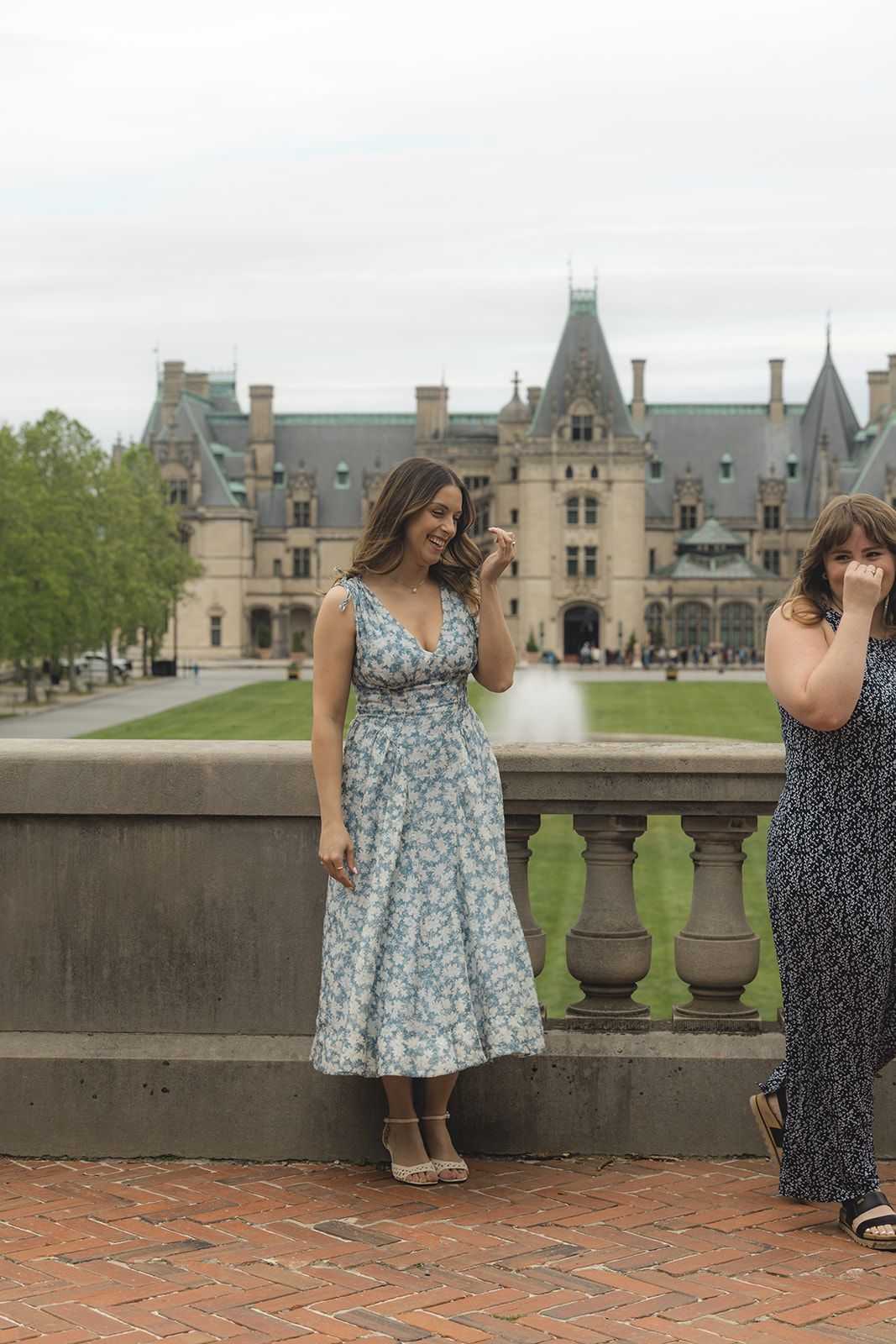 Two women stand on a brick terrace in front of the grand Biltmore Estate, one in a floral dress holding a phone.
