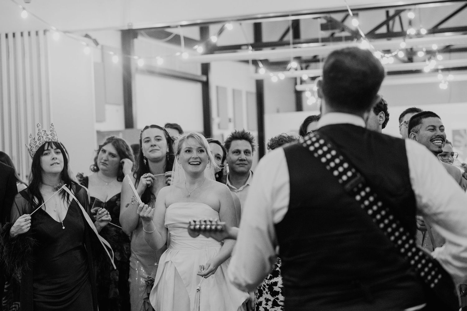 Black and white wedding reception photo of bride smiling at bagpiper performing for guests under string lights.