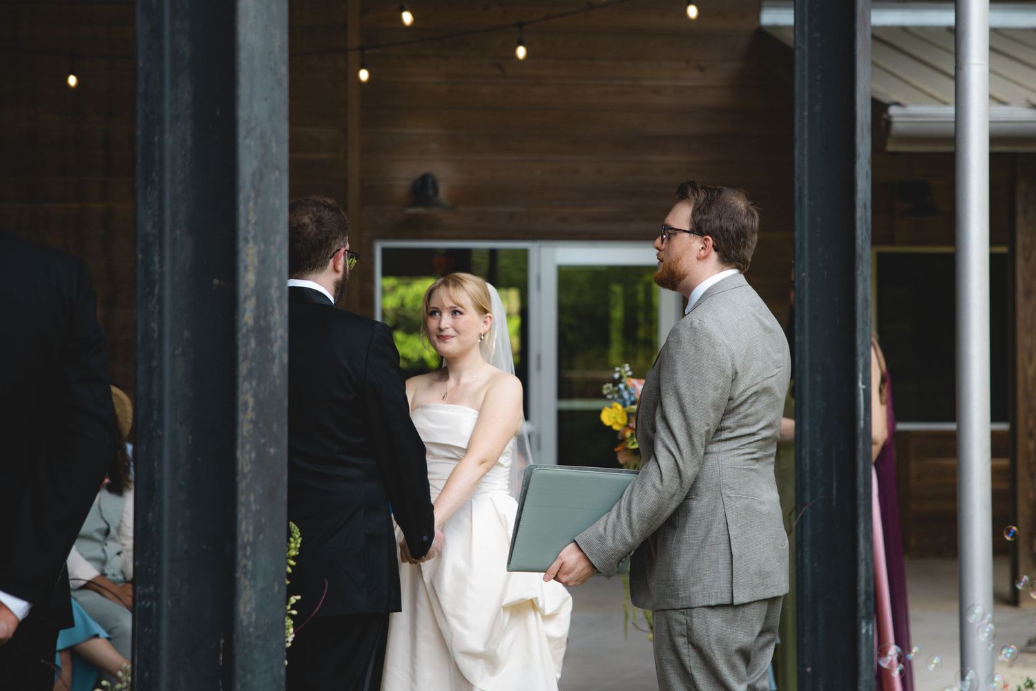 Bride in white gown holds hands with groom in gray suit during outdoor wedding ceremony at rustic venue.
