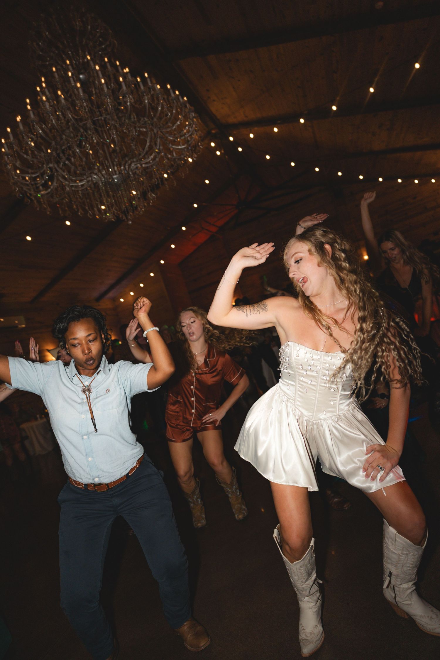 Guests dancing energetically at a rustic barn wedding reception, bride in white dress and cowboy boots.
