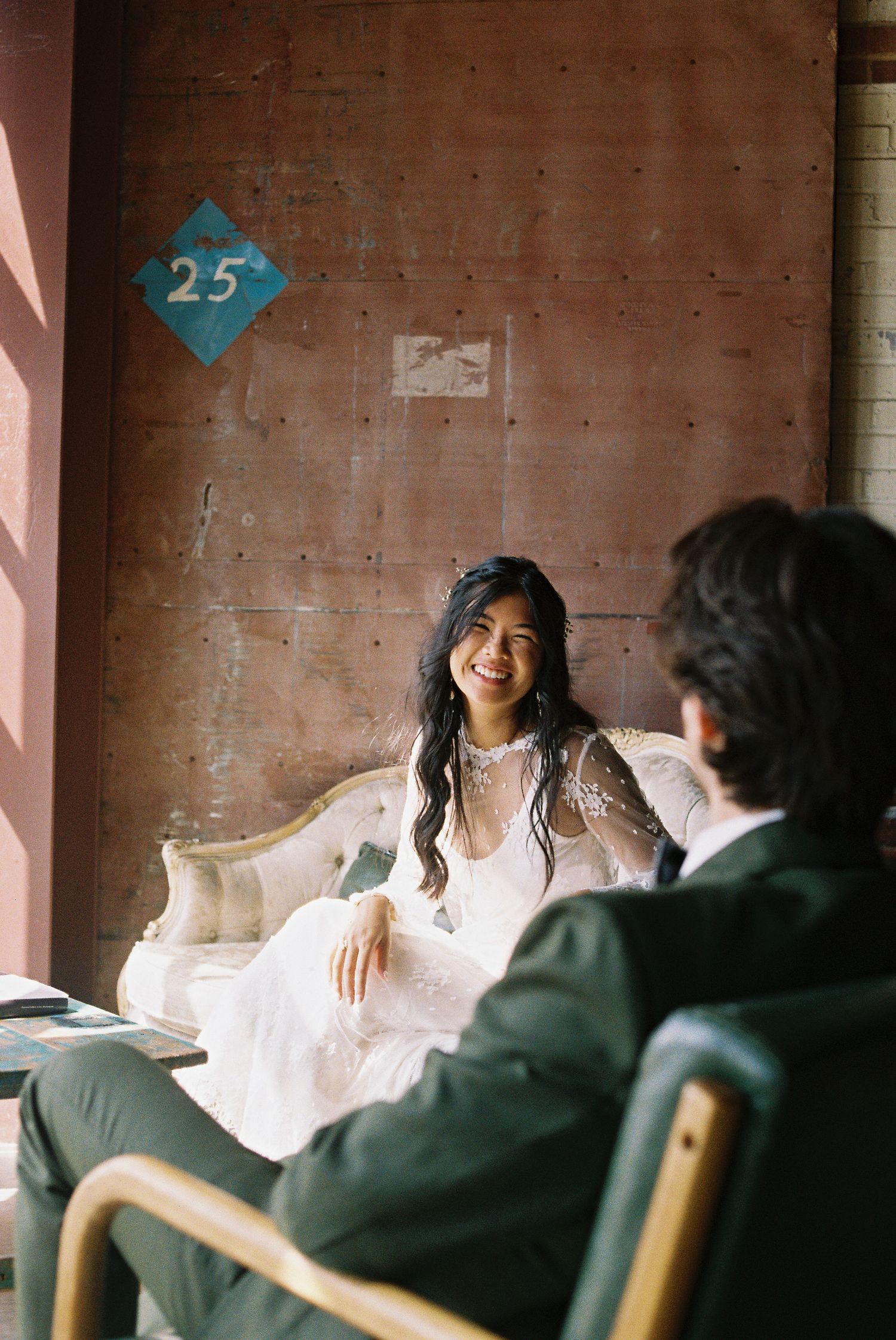 Bride in white lace gown smiles on vintage sofa, groom in green suit seated nearby, industrial venue backdrop.