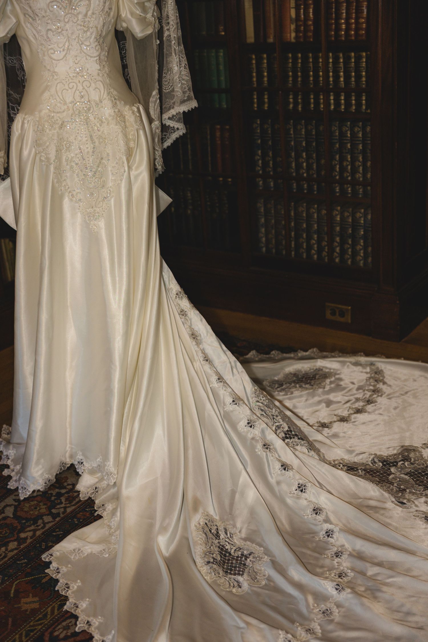 Elegant ivory satin wedding gown with lace trim and long cathedral train displayed in a vintage library setting.