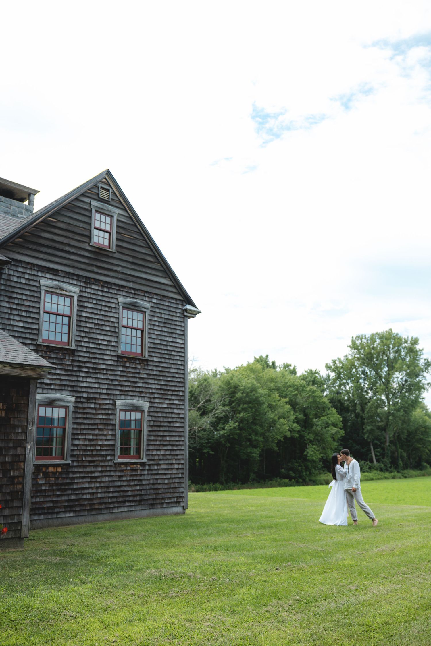 Bride and groom embrace on green lawn beside rustic weathered shingle farmhouse with trees in background.