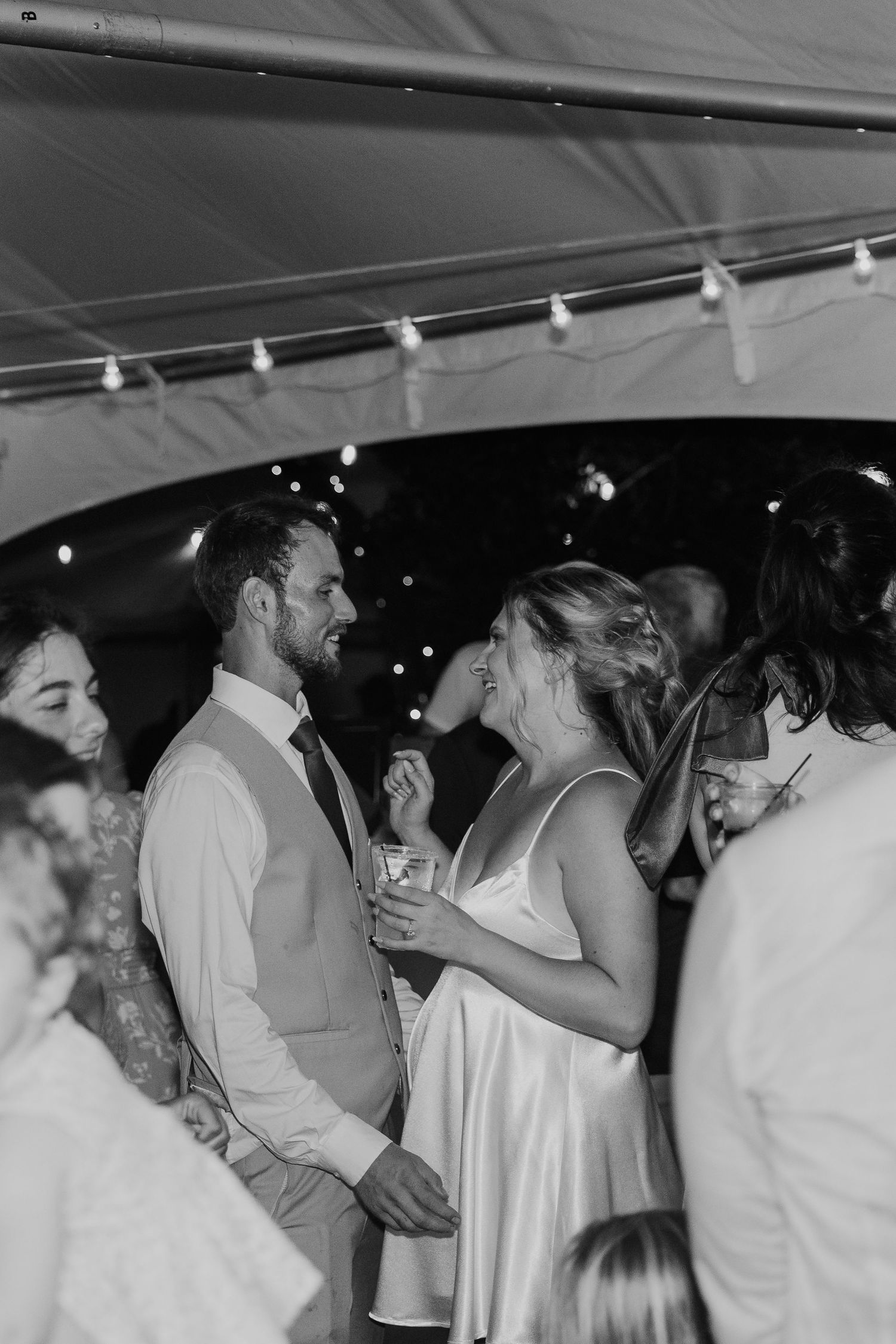 Black and white photo of a bride and groom laughing together at their wedding reception under a tent with string lights.