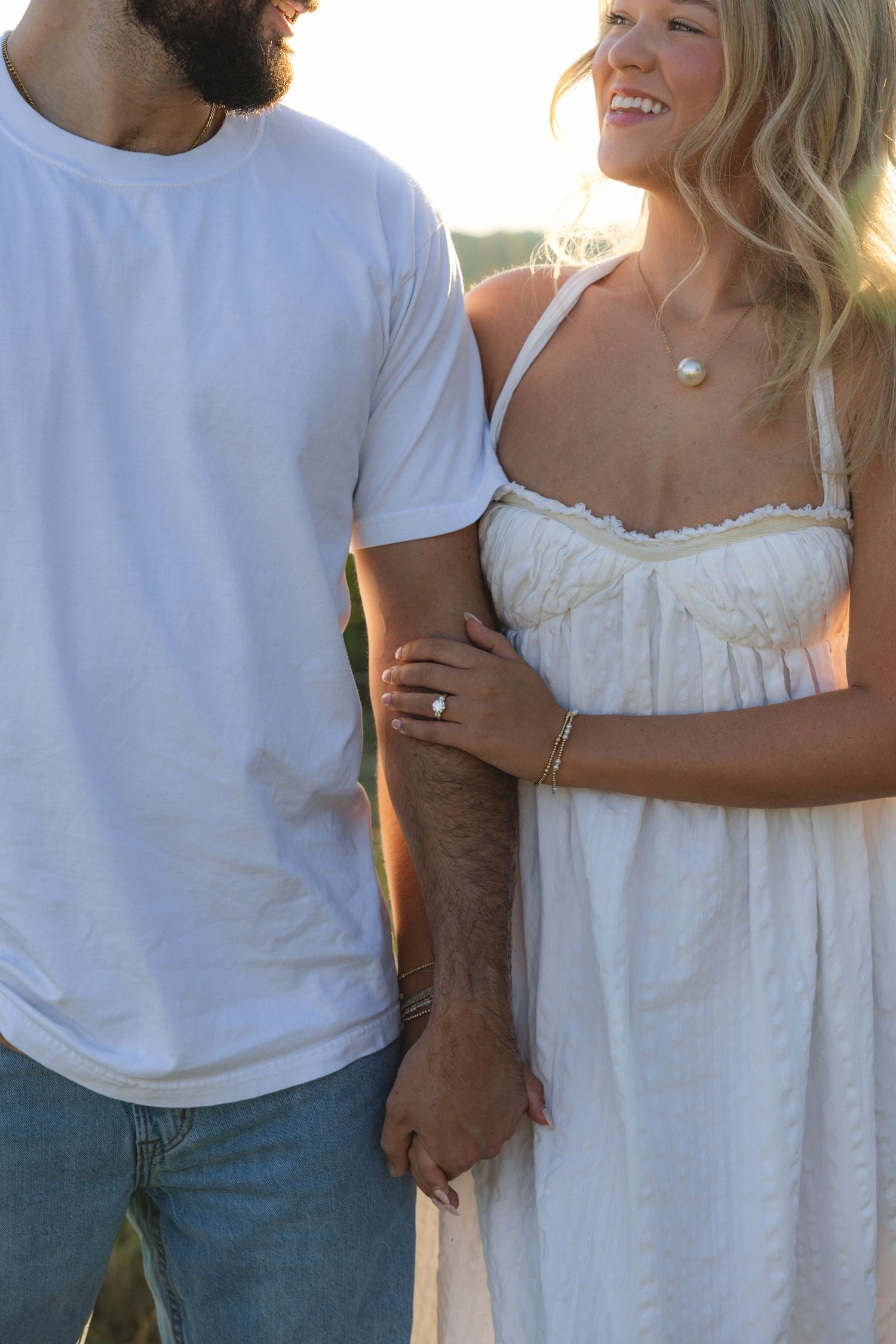 Couple in white outfits holding hands outdoors, woman smiling in sundress with pearl necklace and ring, golden hour light.