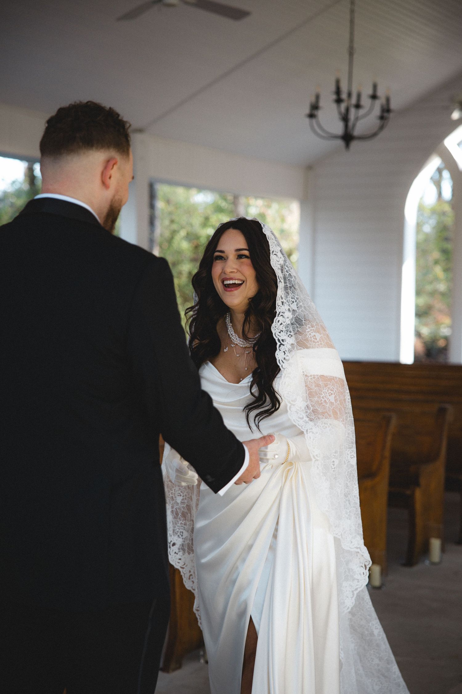 Bride in white lace veil laughing joyfully while holding hands with groom in black suit inside a white chapel.