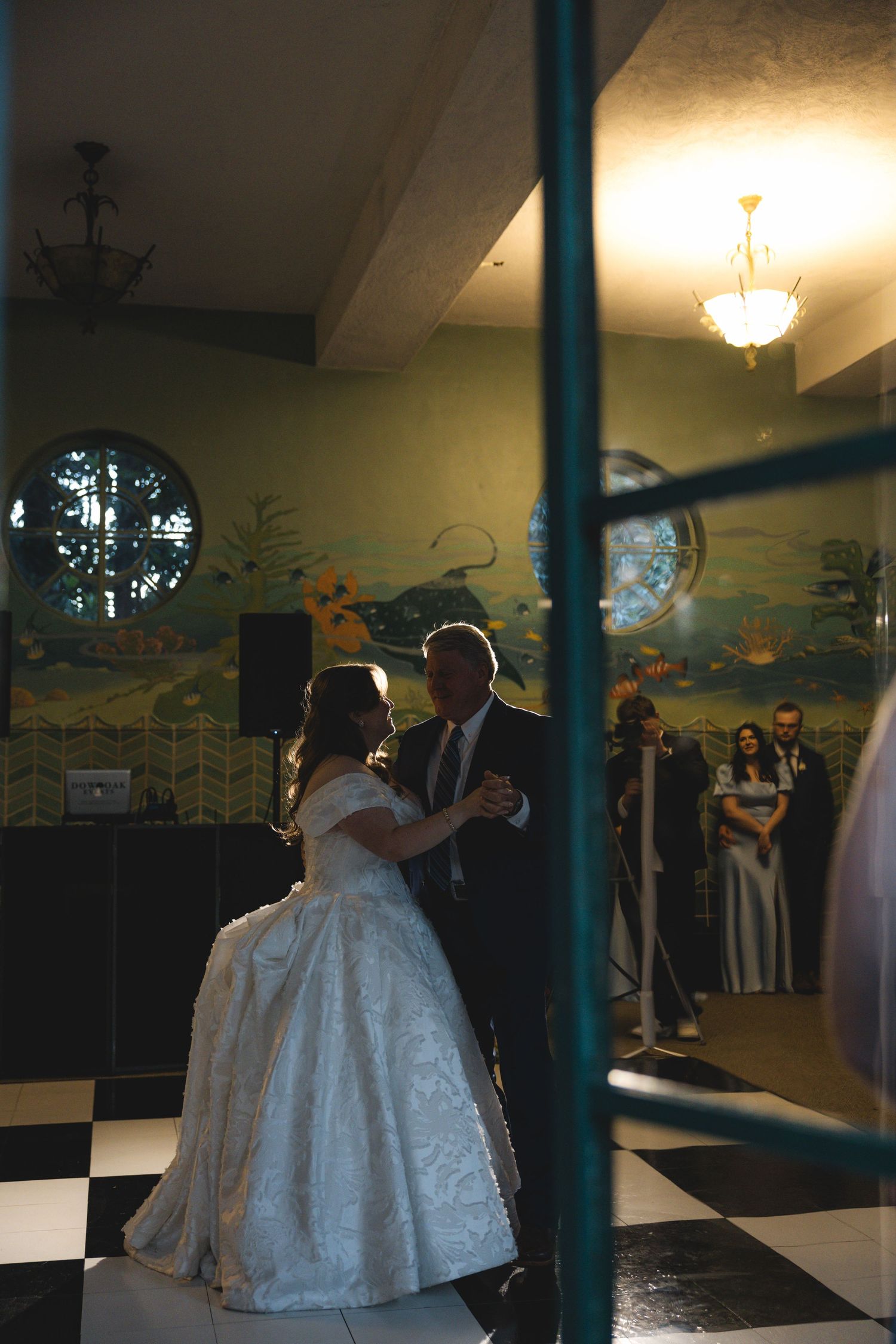 Bride and groom share their first dance on a checkered floor in an elegant venue with murals and guests watching.