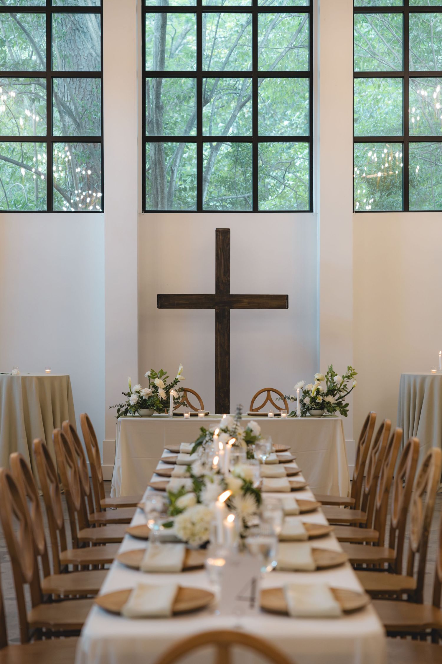 Elegant wedding reception hall with long dining table, wooden cross, white floral arrangements, and large windows overlooking greenery.