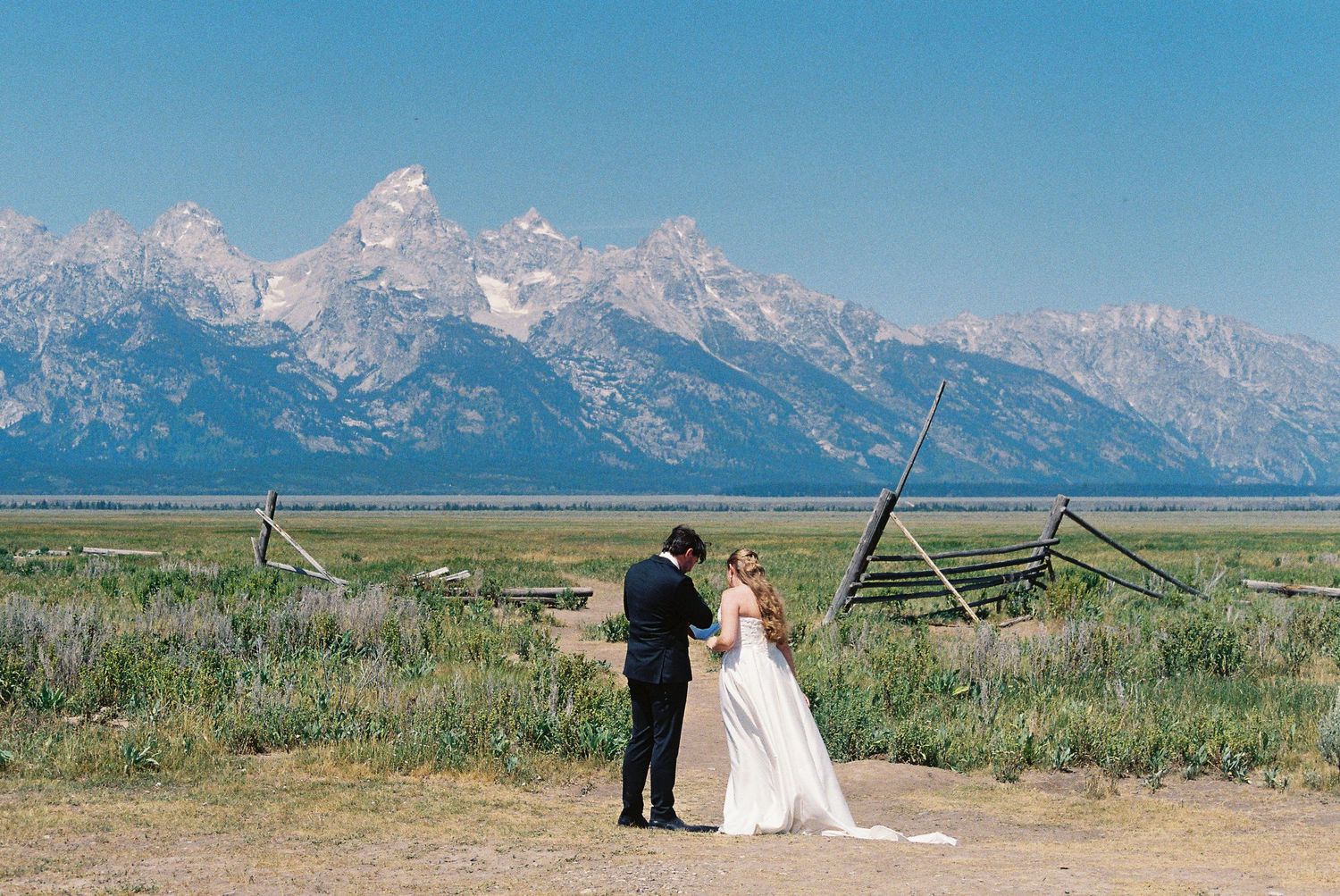 Bride and groom stand before the Grand Teton mountain range in Wyoming, surrounded by open meadows and rustic fencing.