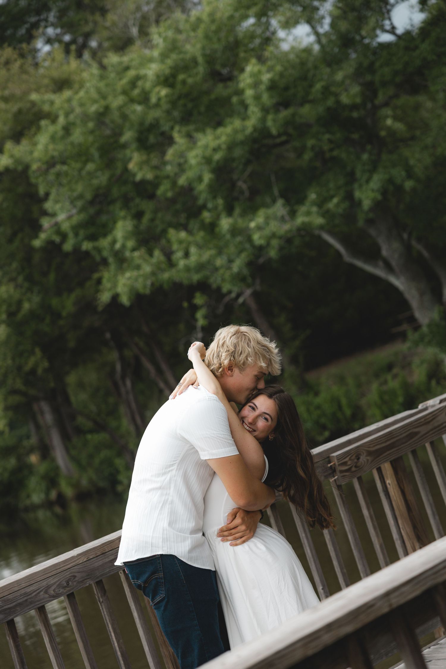 A couple embraces tenderly on a wooden bridge surrounded by lush green trees near a serene waterway.