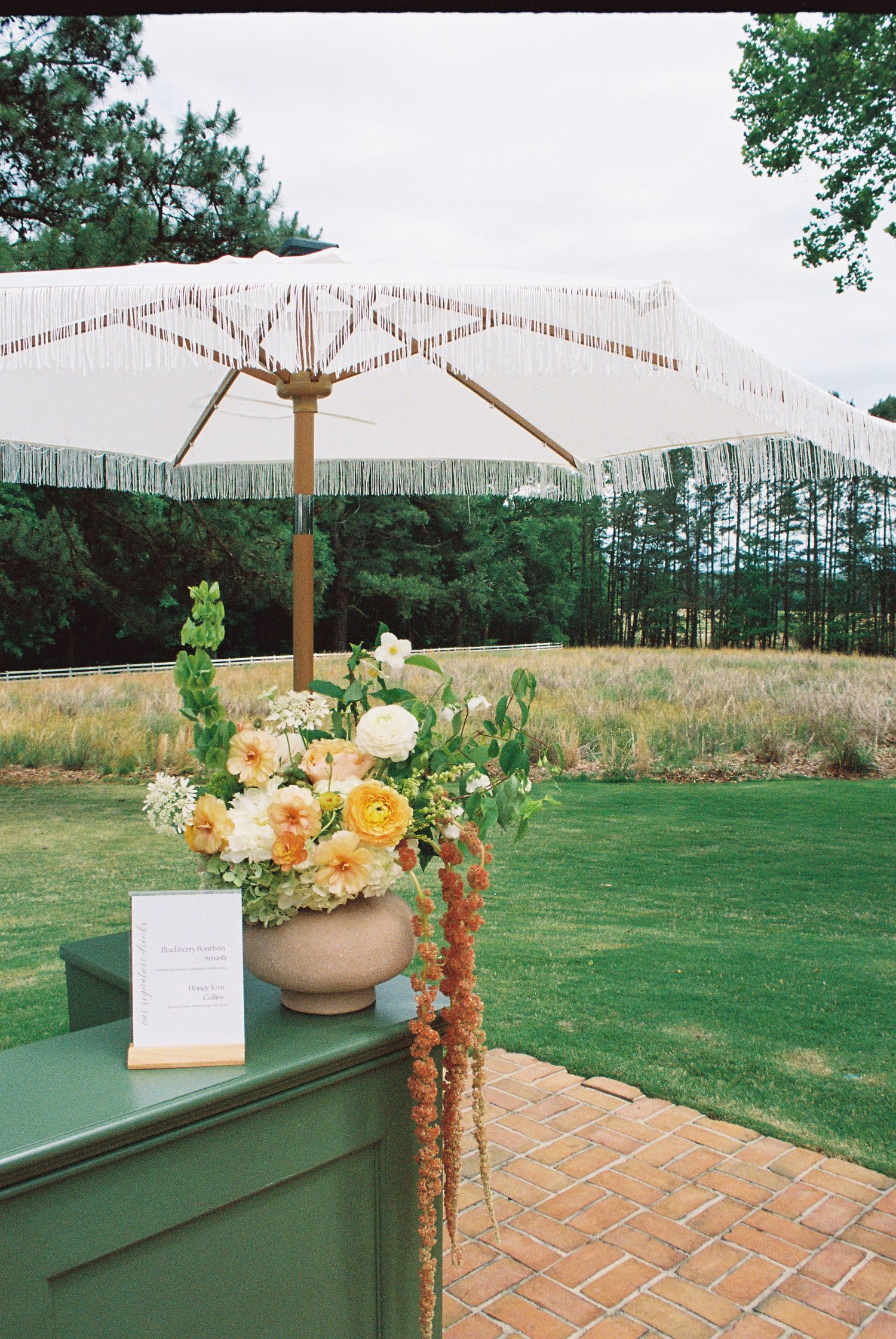 Elegant outdoor wedding entrance with fringed umbrella and yellow floral arrangement on green podium.