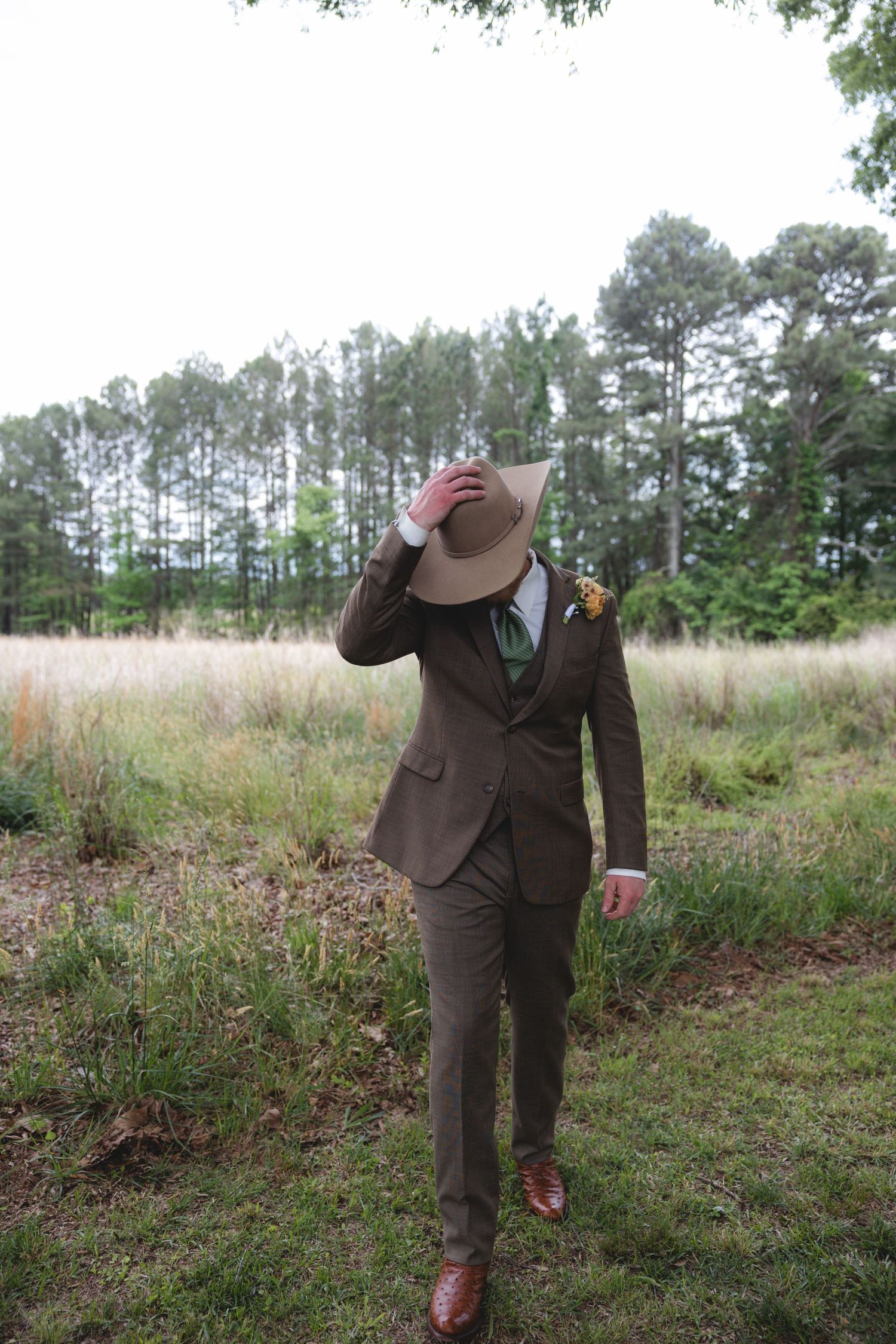 Groom in brown three-piece suit tips cowboy hat while walking through open field.