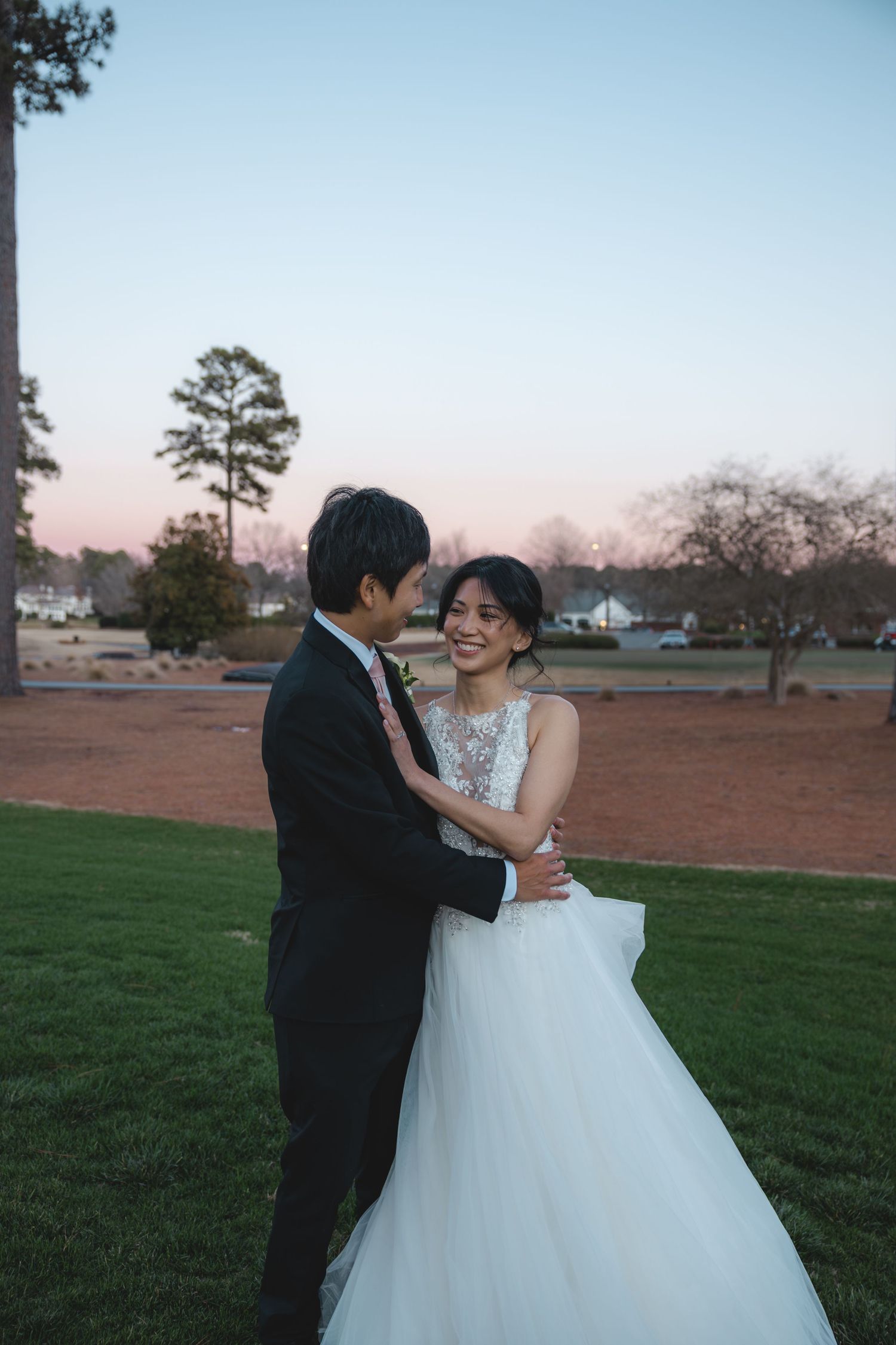 Bride and groom embracing on green lawn at sunset during outdoor wedding portrait.