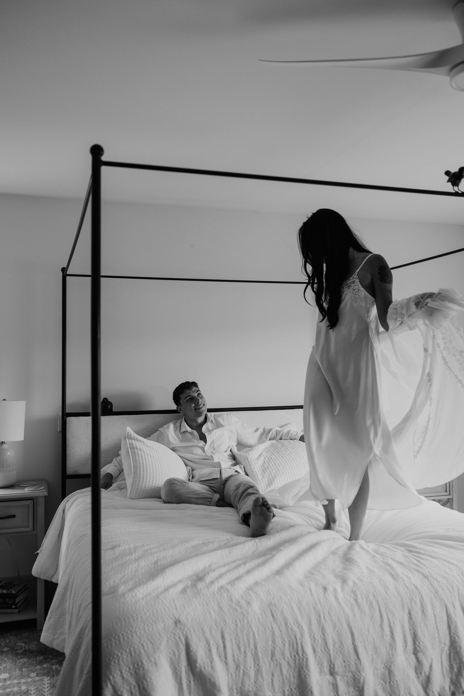 Black and white photo of a couple on a four-poster bed, woman in white dress standing while man sits smiling.
