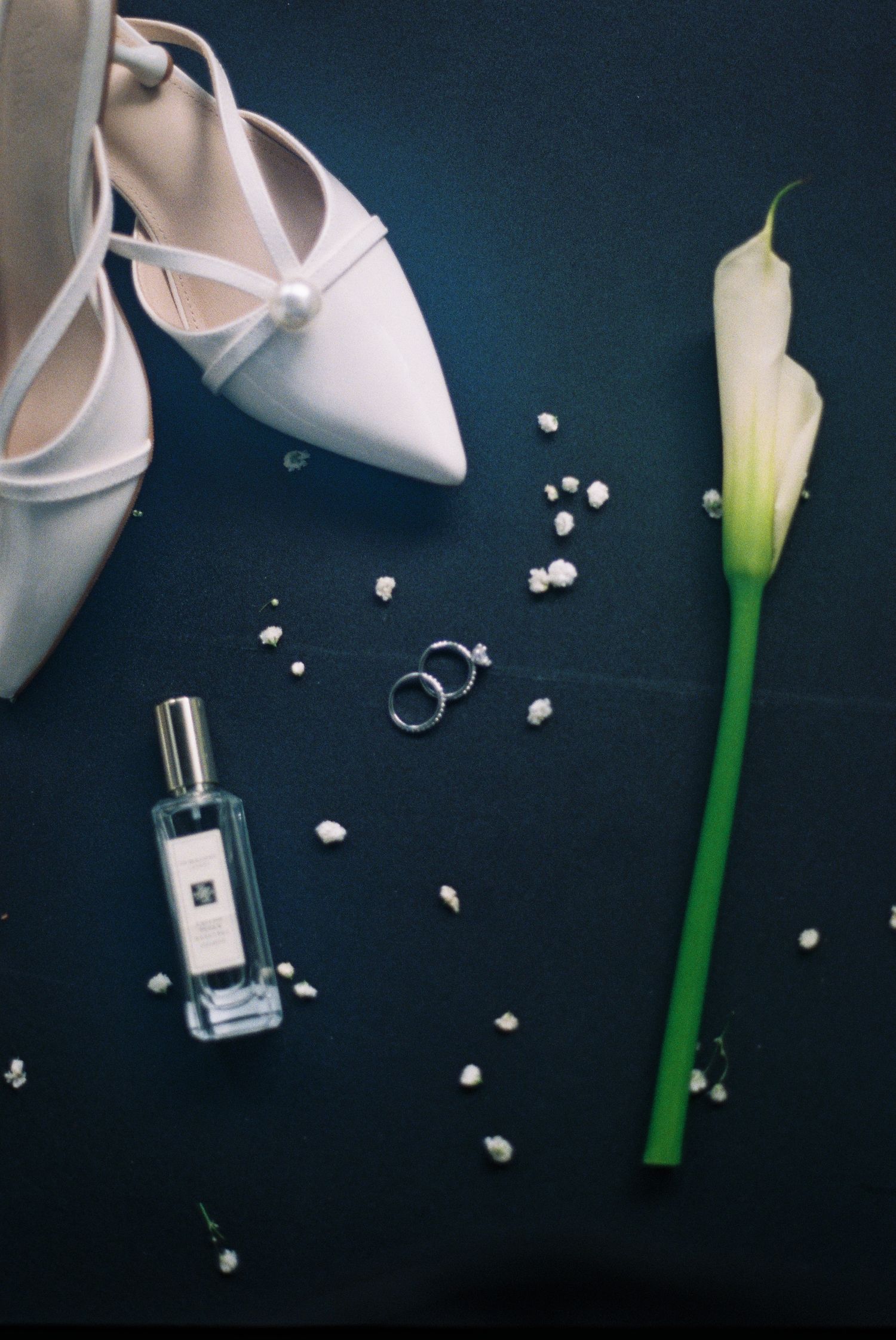 Elegant wedding flat lay with white heels, calla lily, Jo Malone perfume, and rings on dark background.