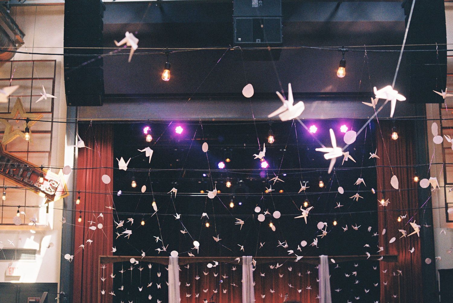 Decorative origami birds and paper ornaments hanging from ceiling with string lights and purple stage lighting.