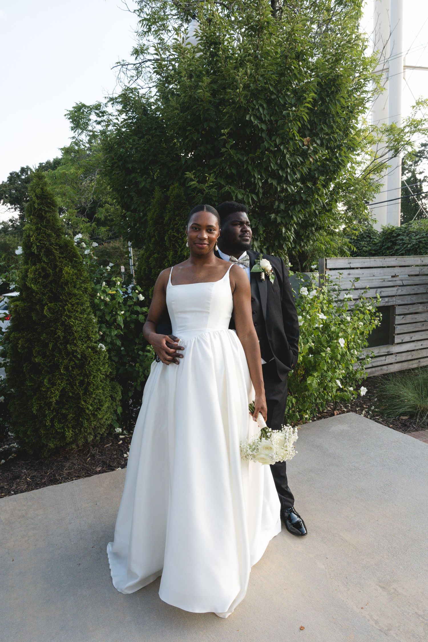 A couple in formal wedding attire poses outdoors, the bride in a white gown holding flowers, groom in a dark suit.