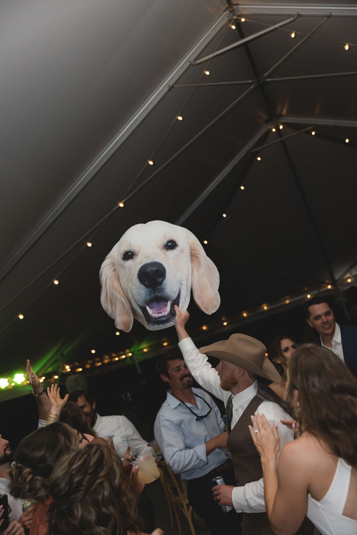 A large golden retriever face balloon held up at a lively wedding reception under a tent with string lights.