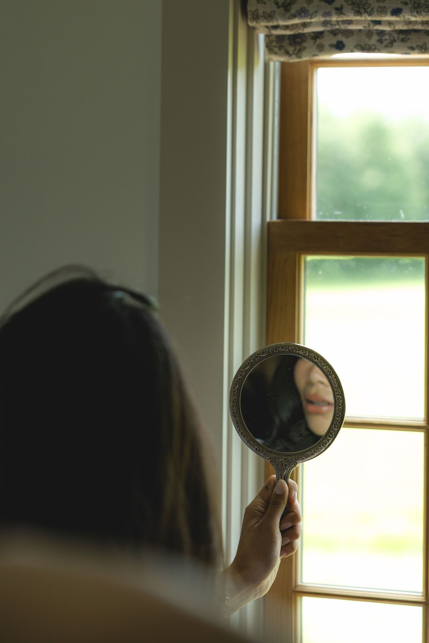 Woman holding ornate hand mirror reflecting her face near a sunlit window with green outdoor view.