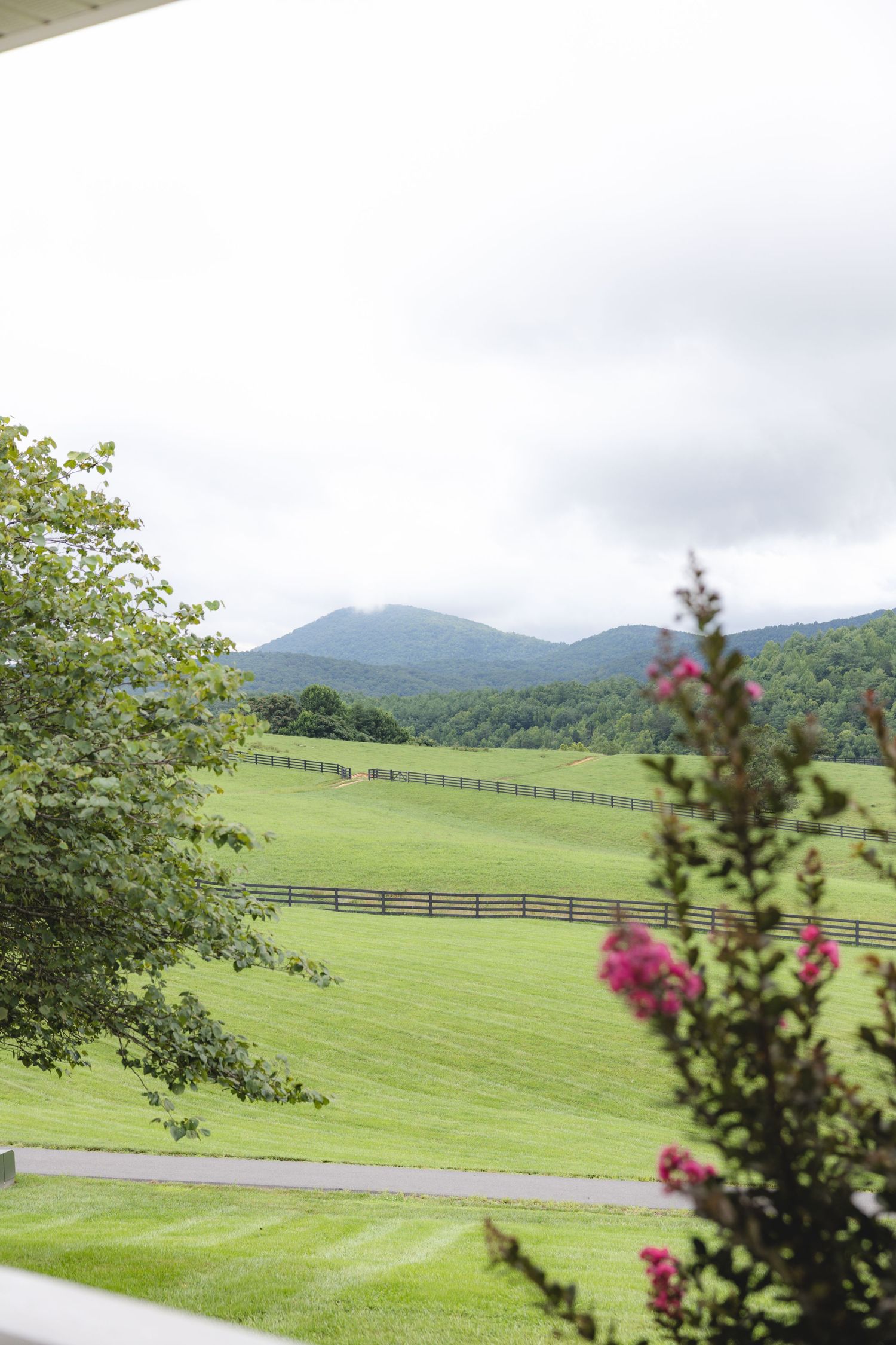 Lush green pastures with wooden fencing, rolling mountains, and cloudy skies viewed from a porch in rural Virginia.